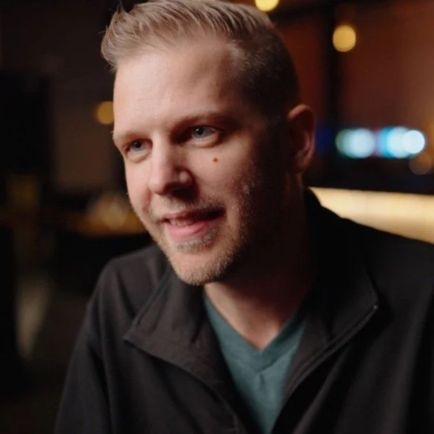 A man with short blond hair and a beard, smiling and looking to the side, wearing a dark jacket and a teal shirt, in a dimly lit indoor setting.