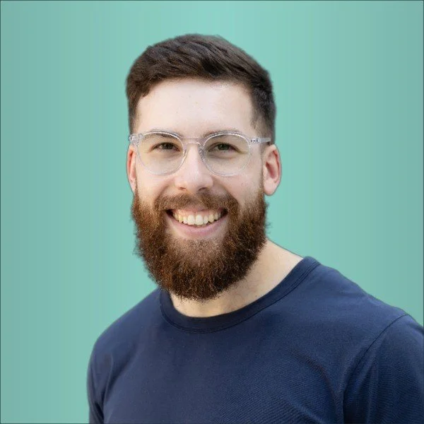 A smiling man with a beard, glasses, and short hair wearing a navy blue shirt, standing against a light teal background.