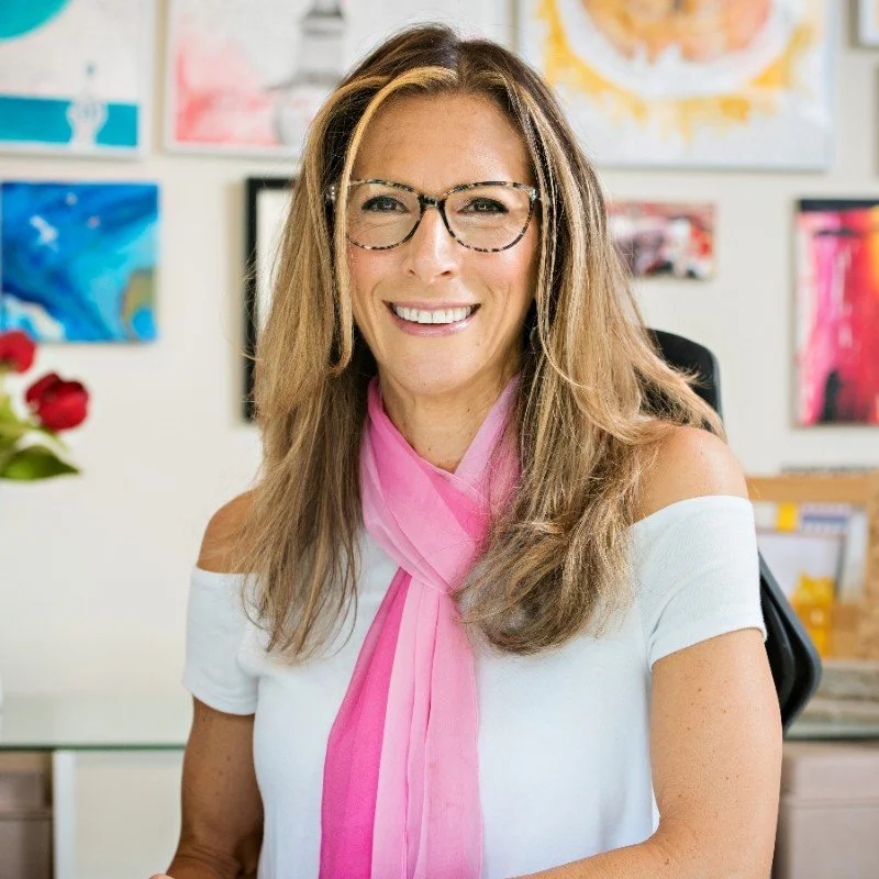 A woman with long hair, glasses, and a bright smile wearing a white off-shoulder top and pink scarf, sitting in an art-filled room.
