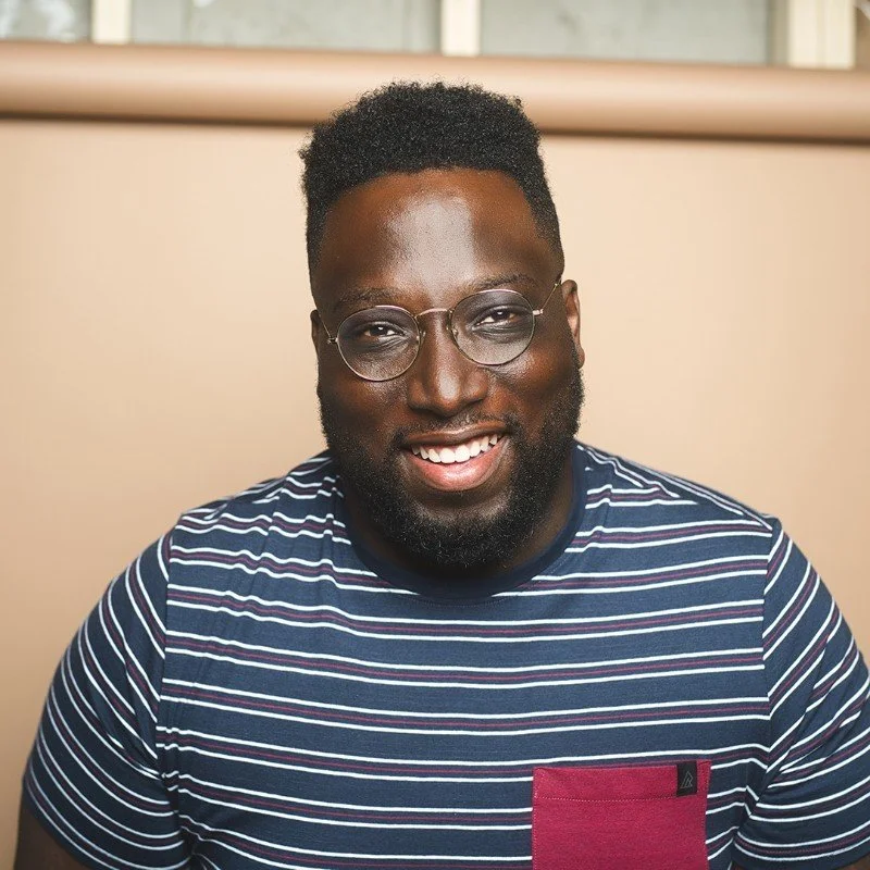 Portrait of a smiling man with dark skin, glasses, a beard, wearing a striped navy and white t-shirt with a maroon pocket, indoors with a beige background.
