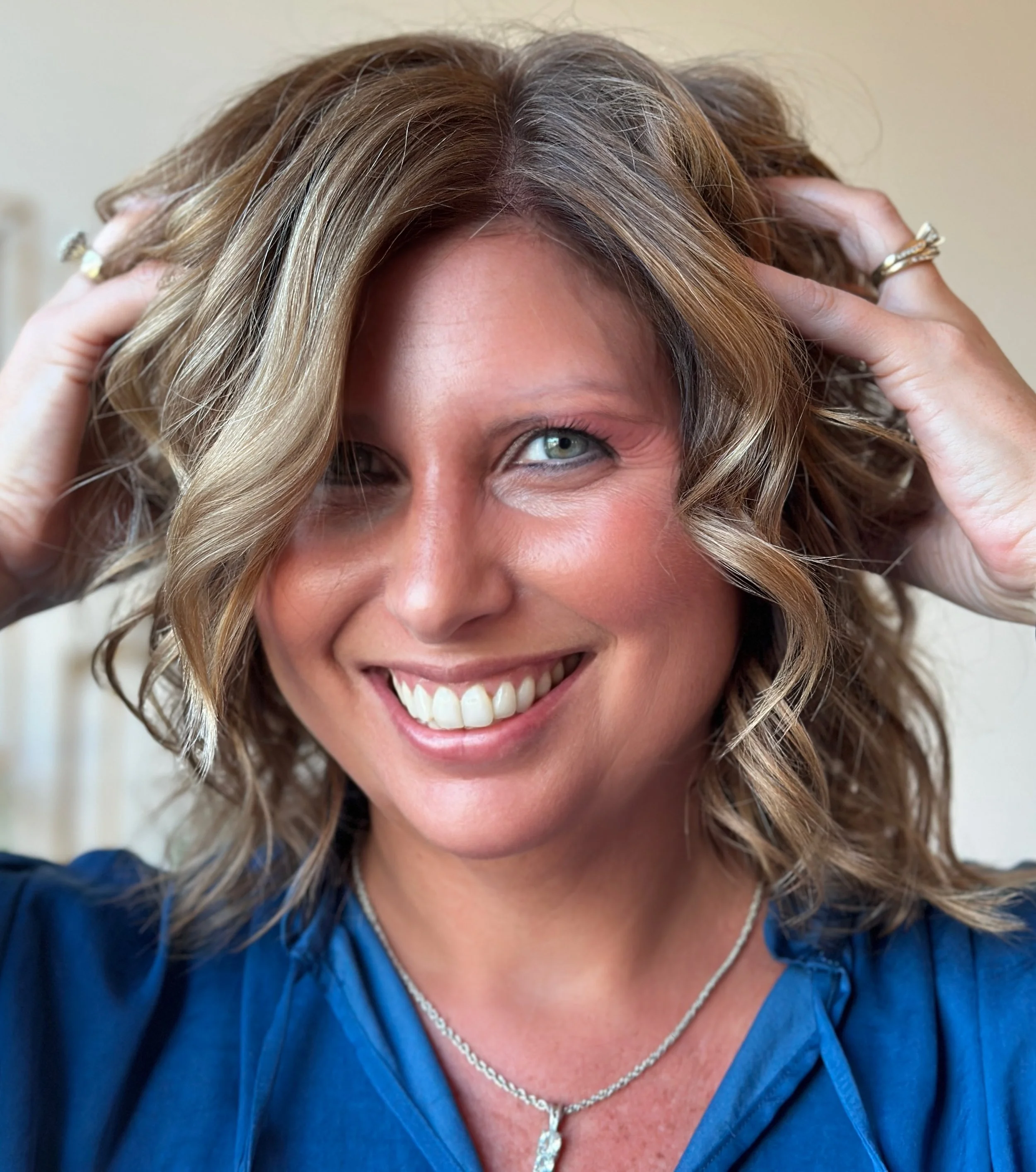 Smiling person with curly hair touching their head, wearing a blue shirt and necklace.