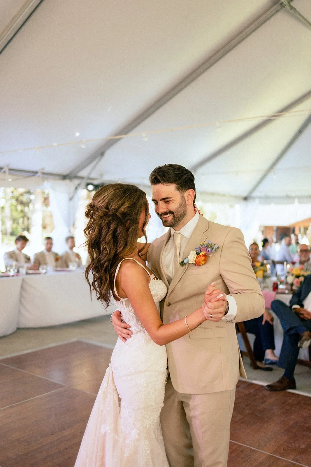 A bride and groom dance closely under a tent during their wedding reception, with guests seated at tables in the background.