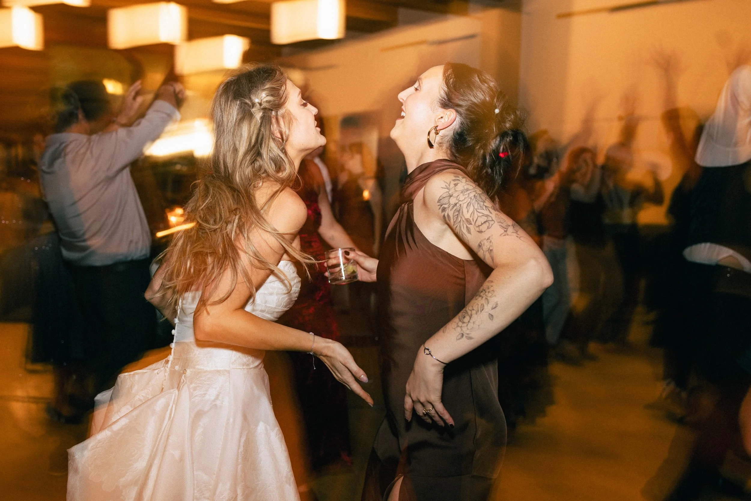 Two women are dancing and smiling at a lively indoor party with warm lighting, surrounded by other guests.