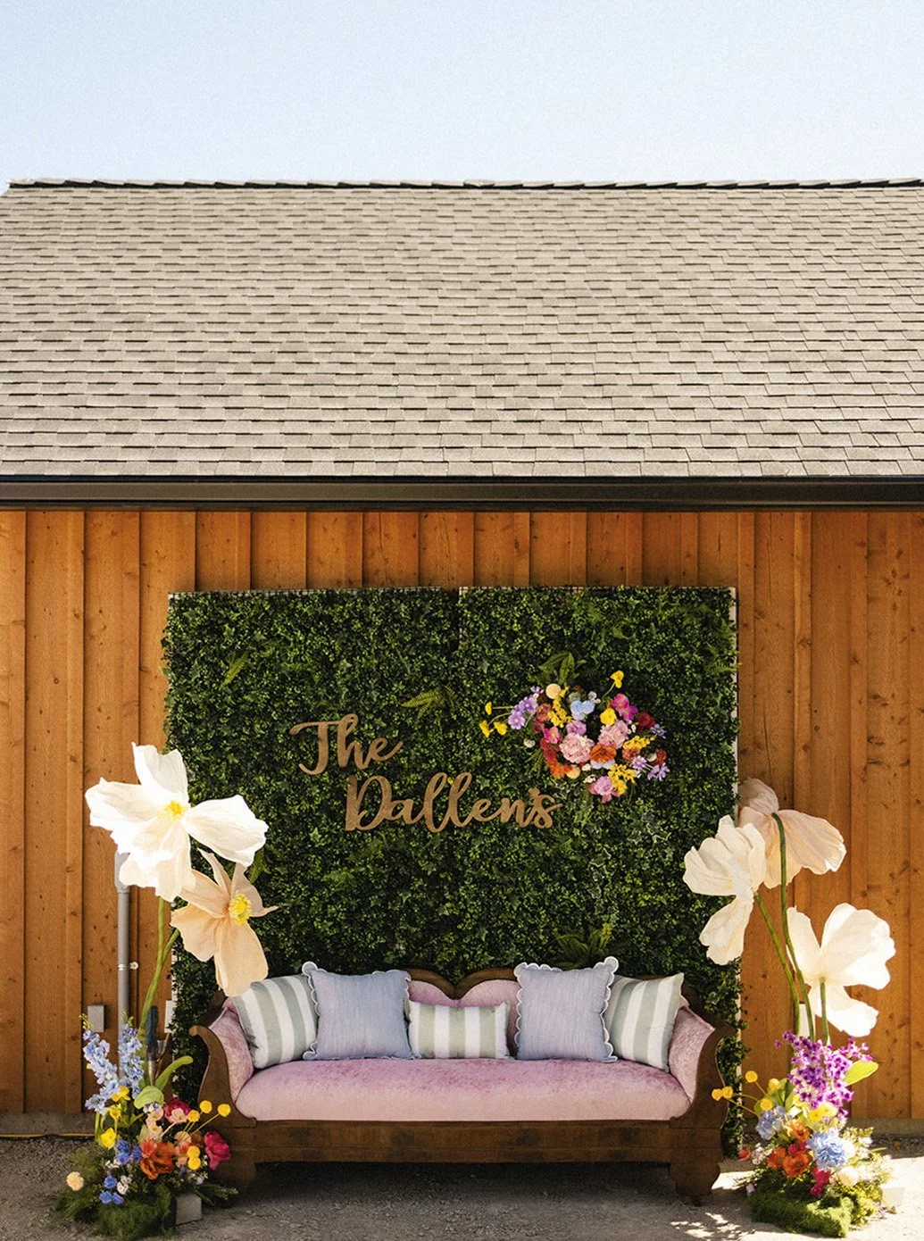 Outdoor decorative display with a wooden wall, green leafy backdrop with the words 'The Dallens,' a pink upholstered sofa with striped and solid pillows, and large white floral arrangements on either side, accented with smaller colorful flowers.