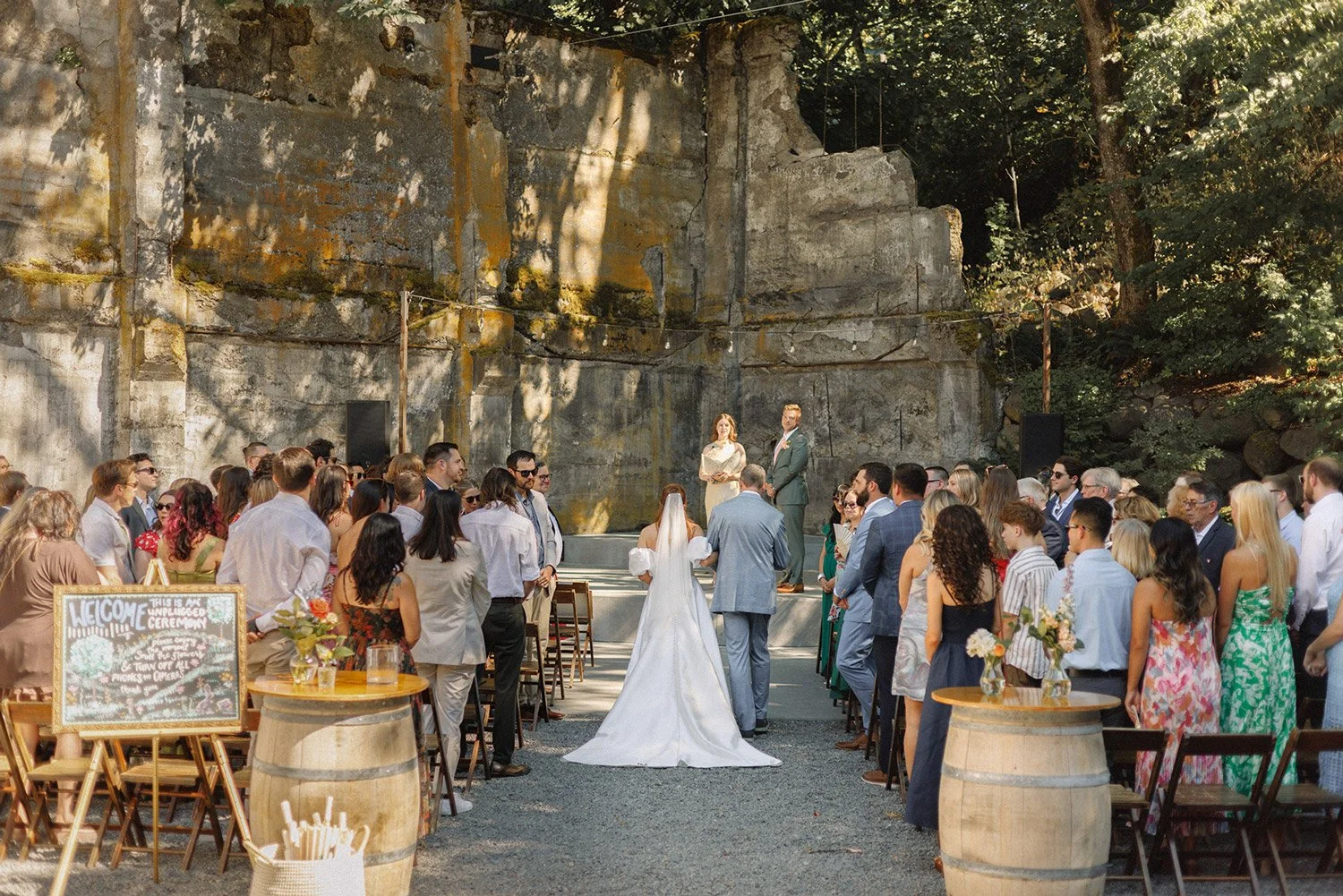 A wedding ceremony taking place outdoors in front of a rocky wall with many guests attending. The bride and groom are standing on a small stage with the bride facing away, dressed in white, and the groom facing forward in a gray suit. Guests are standing on either side of the aisle, some wearing sunglasses, and a welcome sign is visible on a barrel to the left.