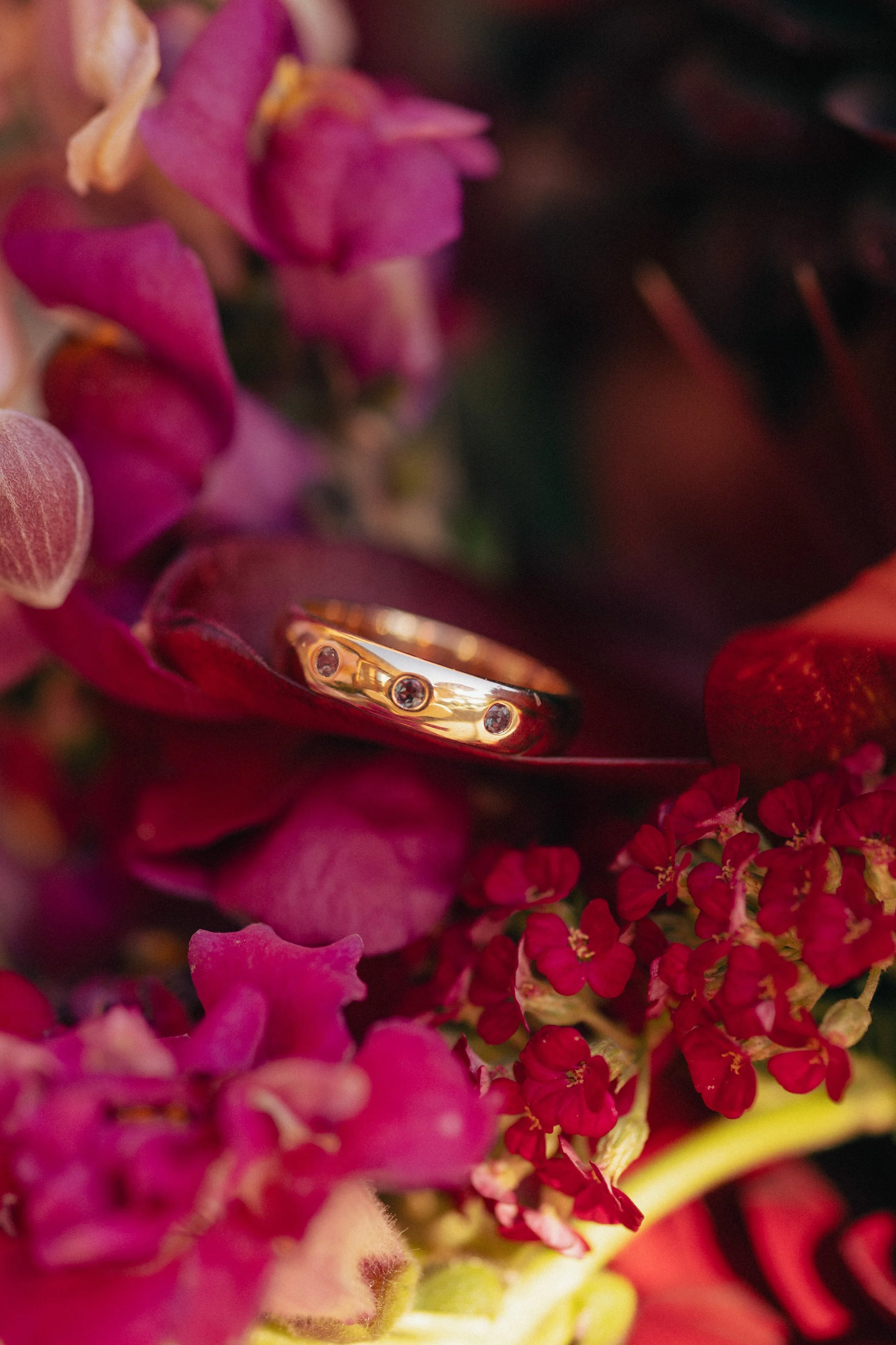 A close-up of a gold ring with embedded diamonds among vibrant pink and red flowers.