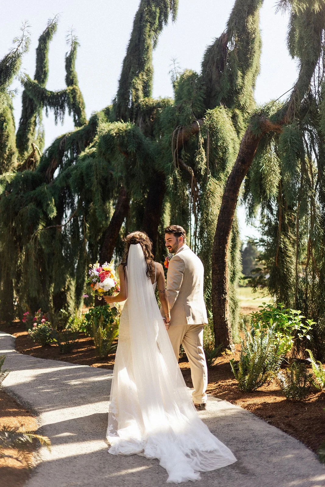 A bride and groom walking hand in hand outdoors under large green trees, with the bride in a white wedding dress holding a colorful bouquet, and the groom in a light beige suit, both smiling.