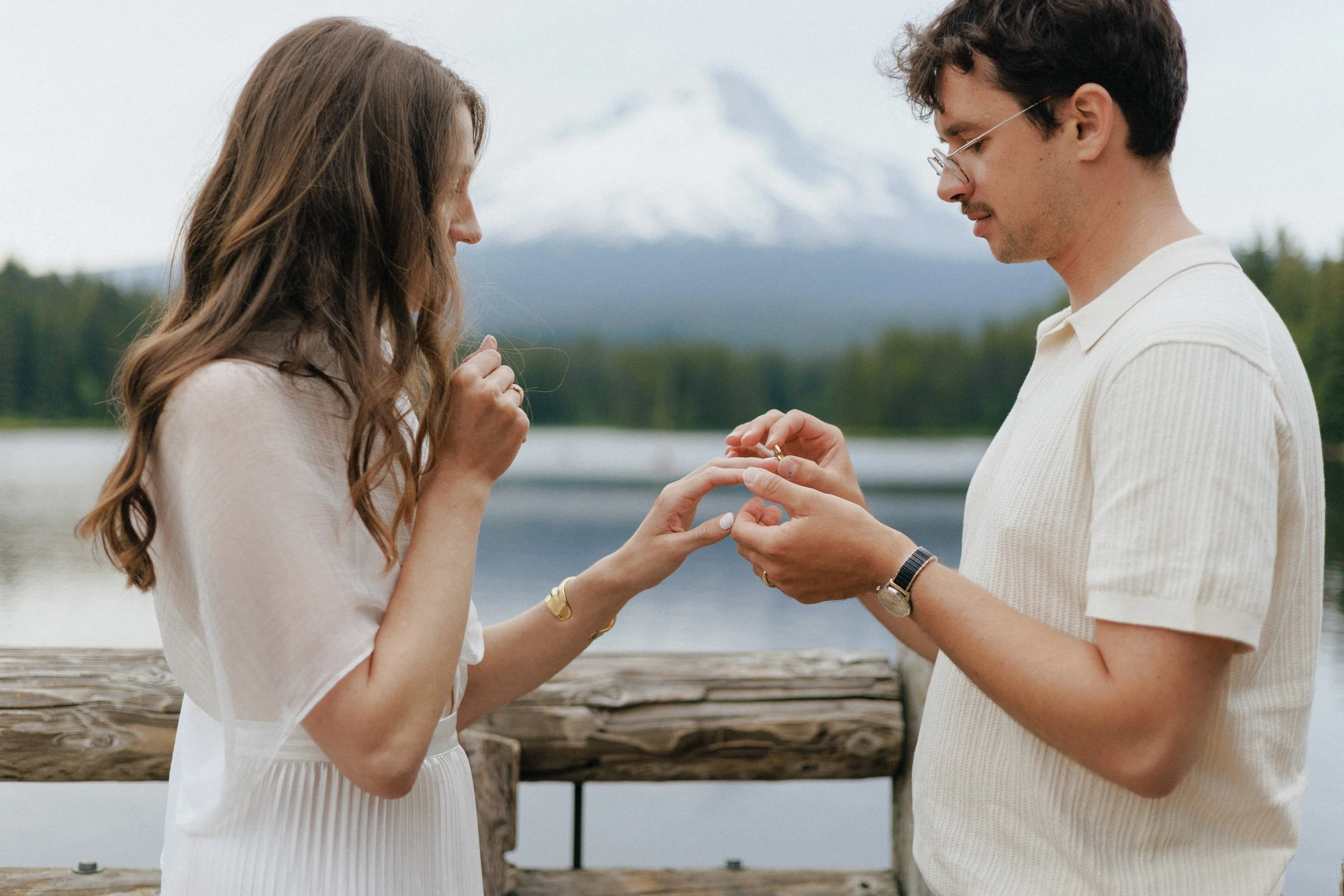 A couple is standing outside by a body of water with mountains in the background, and the man is placing a ring on the woman's finger during a proposal.
