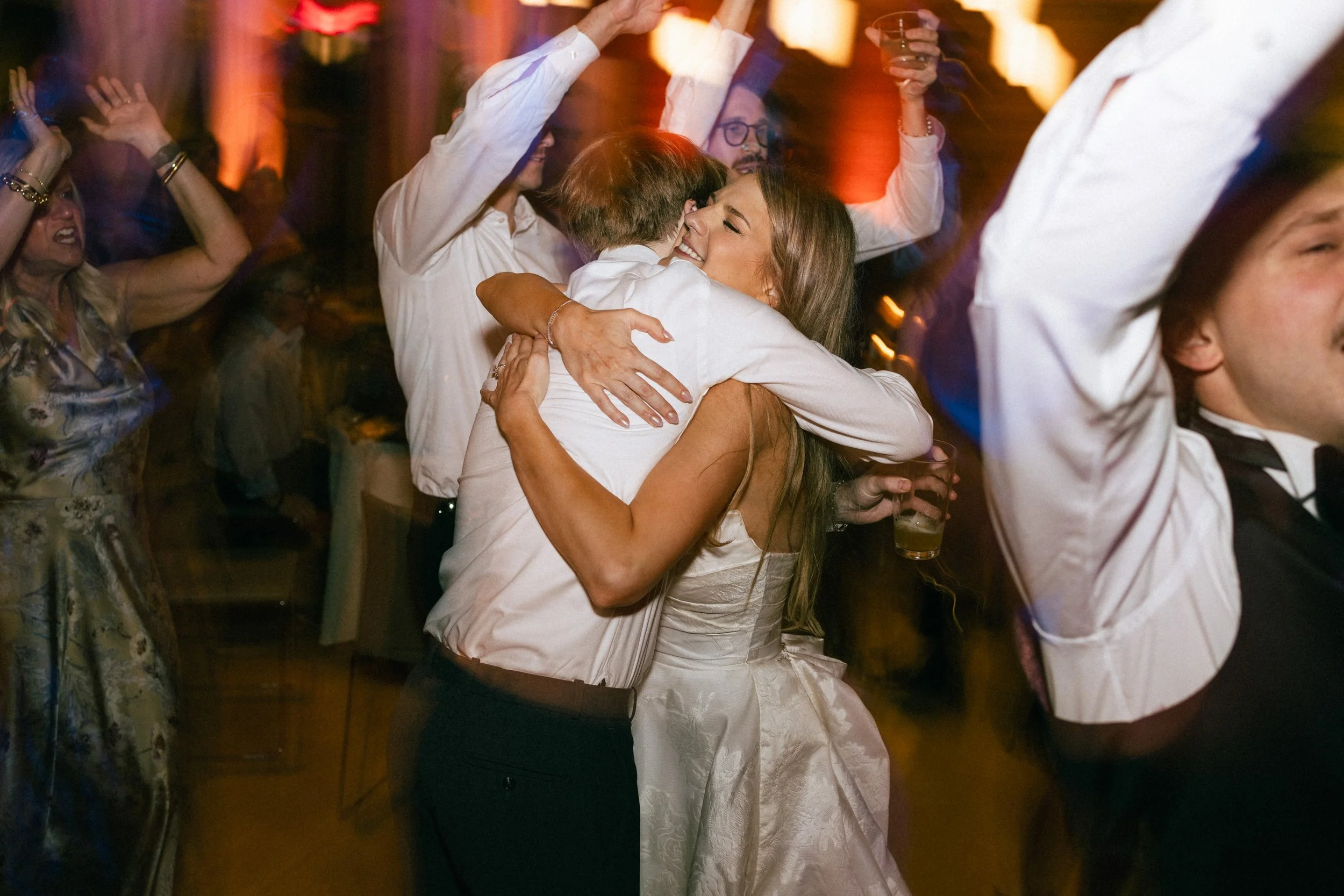 People dancing and hugging at a lively indoor party with warm lighting and drinks in hand.