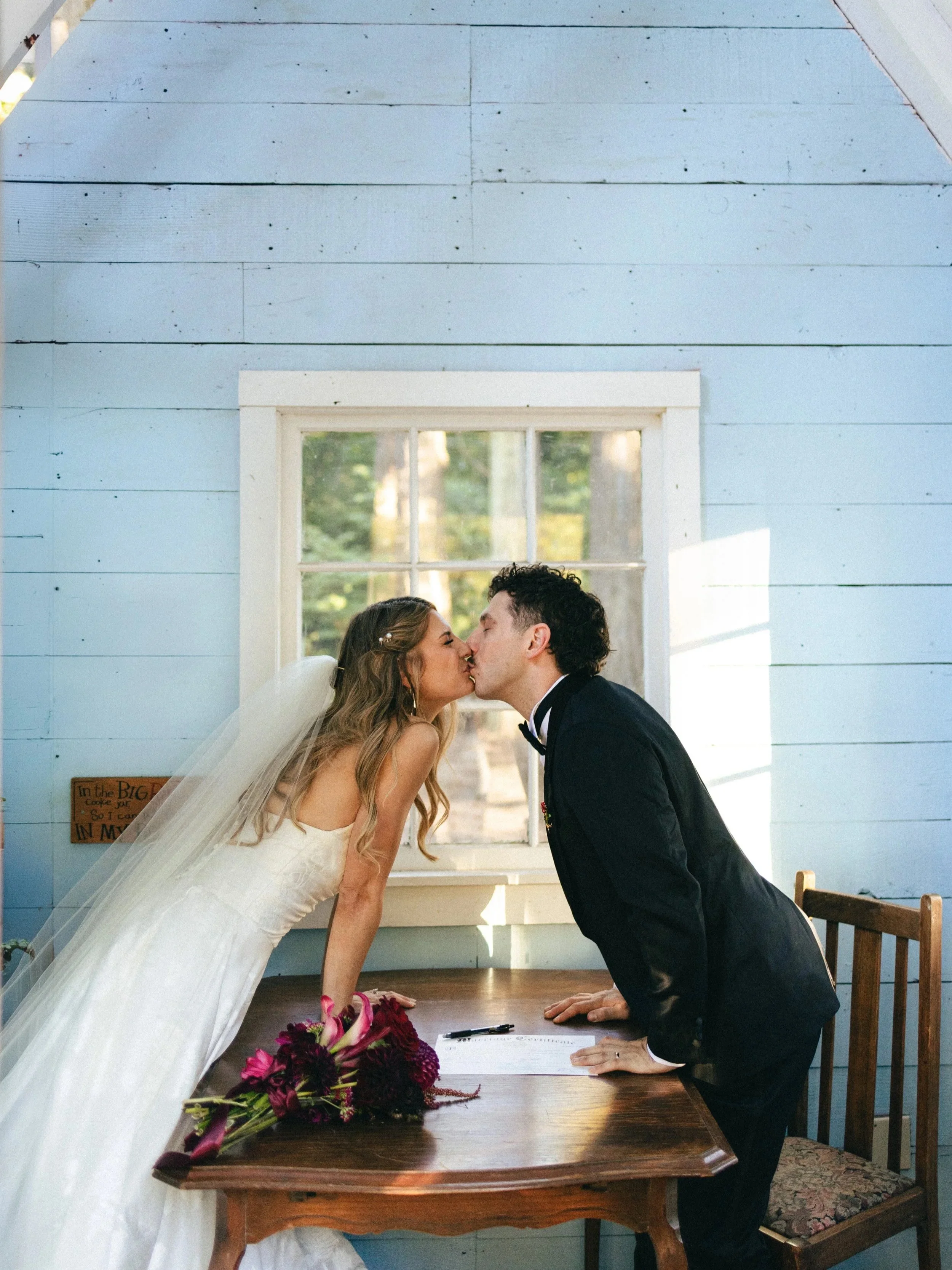A bride and groom are kissing inside a room with blue wooden walls and a window. The bride is wearing a white wedding dress and veil, holding a bouquet of flowers. The groom is in a black tuxedo. They are leaning over a wooden table with a bouquet and a pen on it.