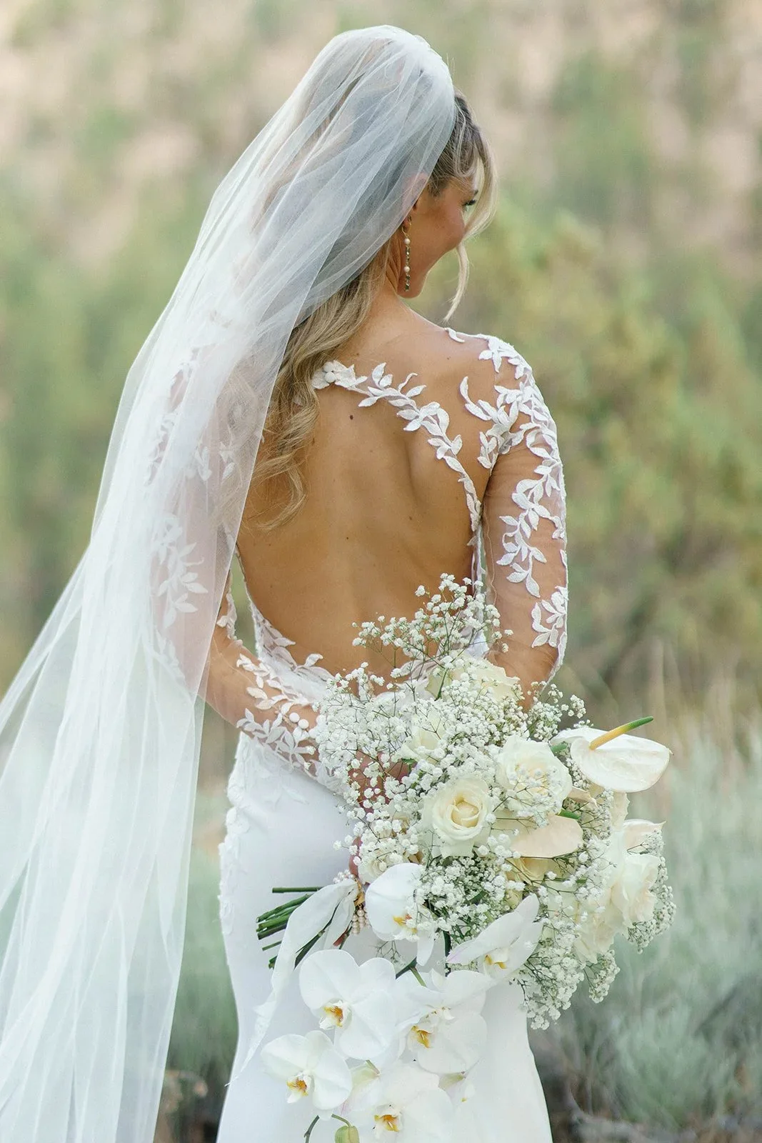 Bride in a wedding dress with a lace back and a long veil, holding a bouquet of white flowers, standing outdoors.
