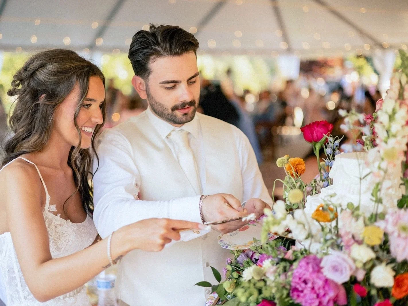 A bride and groom cutting their wedding cake together at a reception with floral decorations and guests in the background.