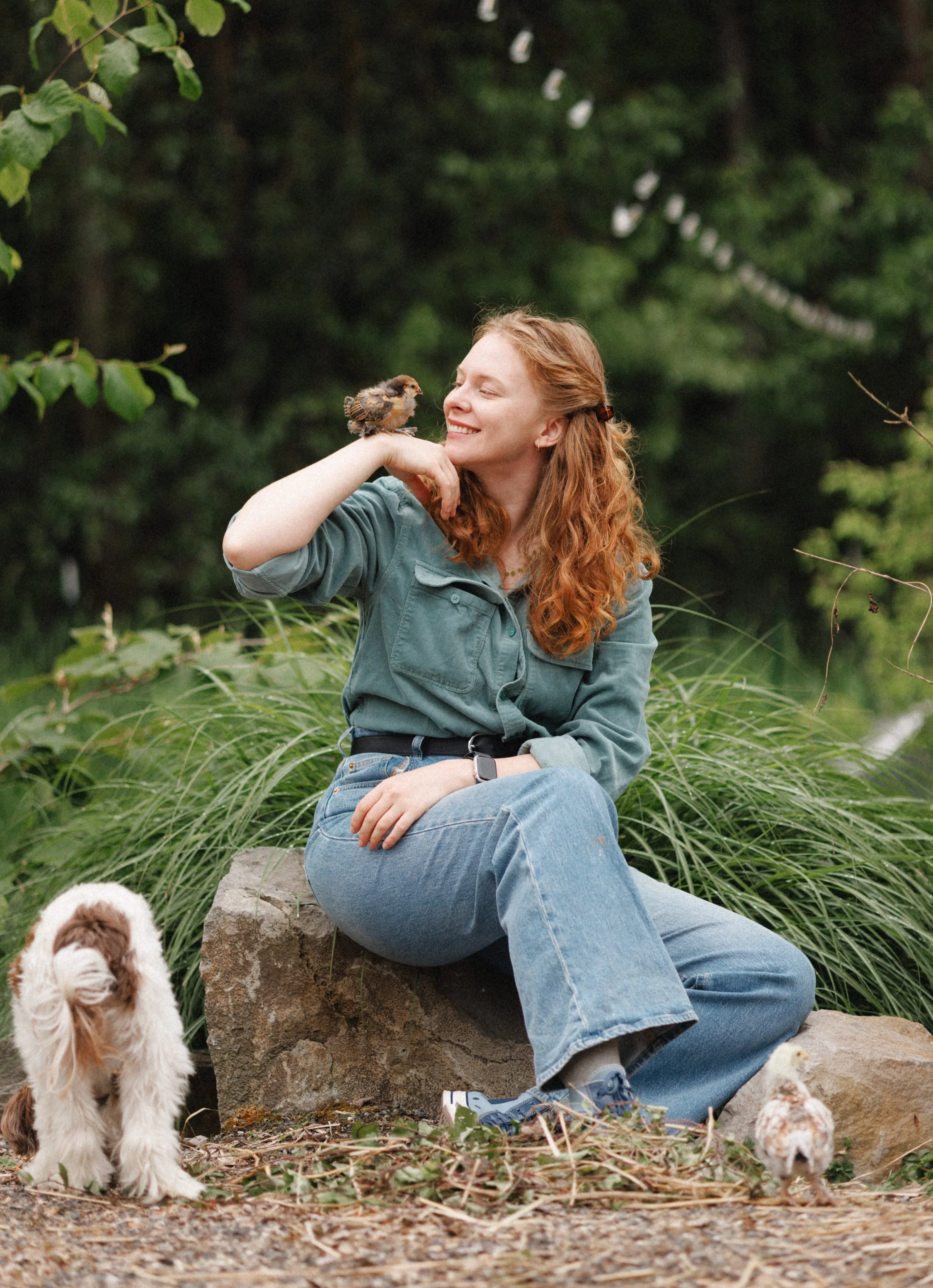 A smiling woman with curly red hair, wearing a teal jacket and blue jeans, sitting on a rock outdoors in a green forested area. She has a small brown and white baby chick perched on her hand, and there are other chicks at her feet and nearby.