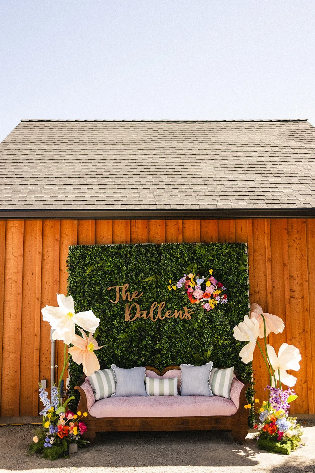 A decorated outdoor space with a wooden wall, green hedge backdrop with a floral arrangement and the words 'The Dallens', a pink vintage sofa with striped cushions, and large white flowers in front.