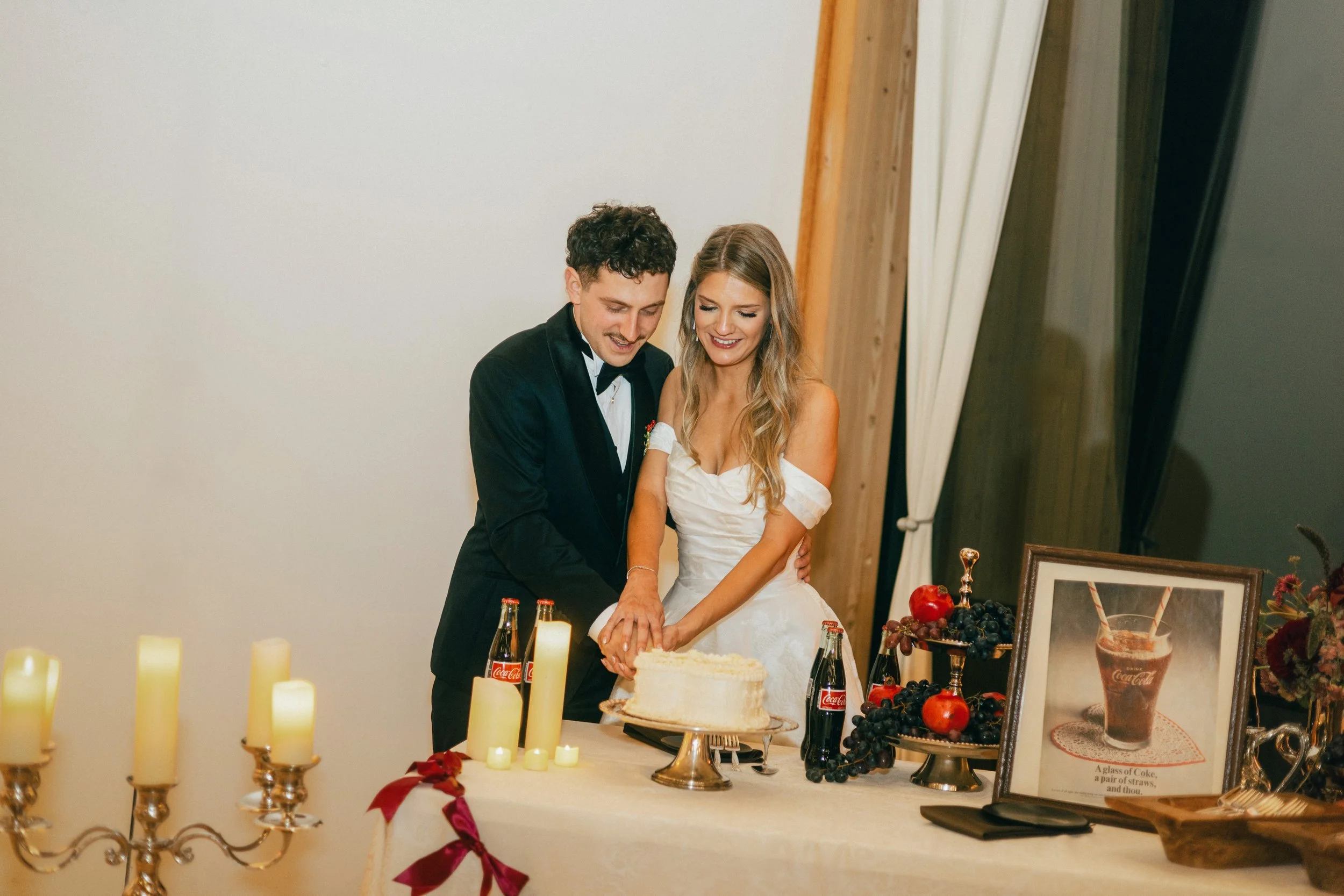 A newlywed couple cutting a wedding cake at their celebration, surrounded by candles, fruit, and framed pictures, in a warmly decorated room.