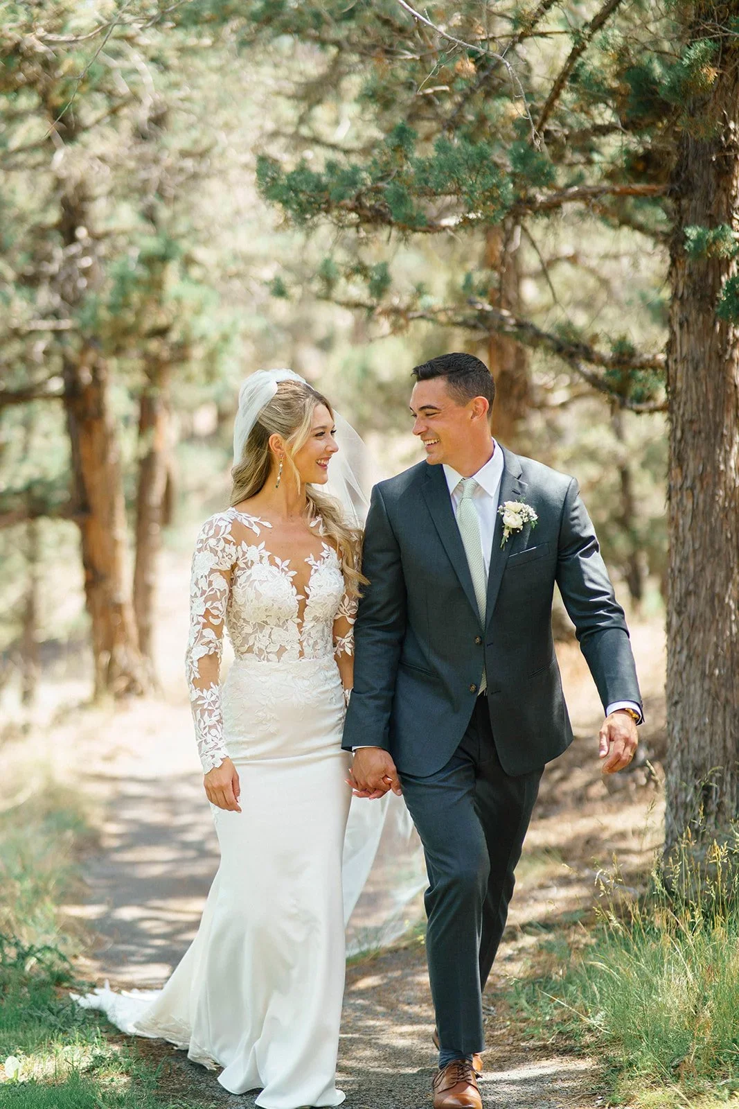 A bride and groom walking hand in hand through a wooded outdoor area, smiling at each other.