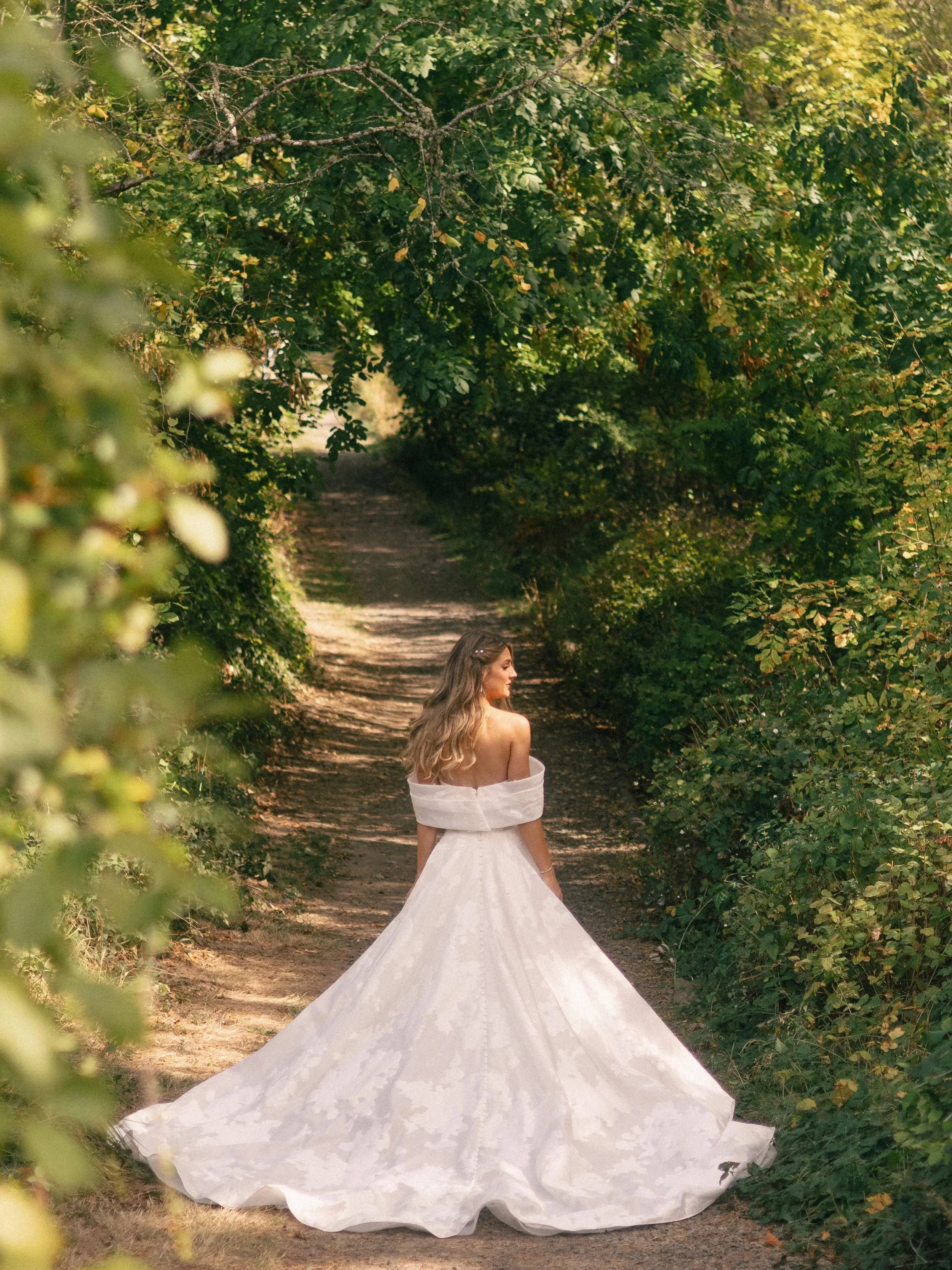 A woman in a white wedding gown standing in a forested path with green trees arching overhead.