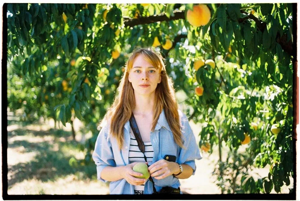 A young woman with long light brown hair standing under a fruit-laden tree in an orchard, holding an apple and a smartphone, wearing a blue shirt over a striped top.