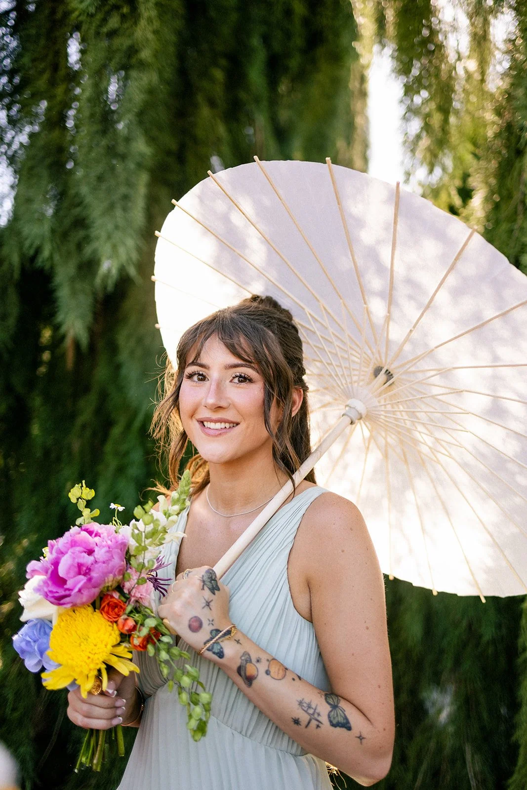 A young woman smiling outdoors holding a bouquet of colorful flowers and a white parasol, with green trees in the background.