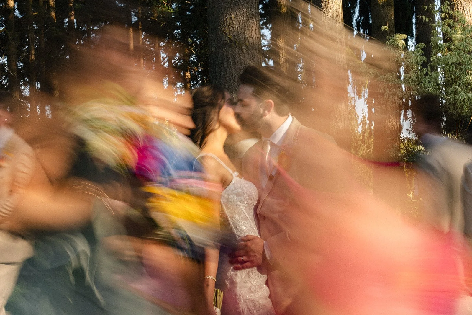 A couple sharing a kiss at an outdoor wedding, surrounded by blurred guests and trees in the background.