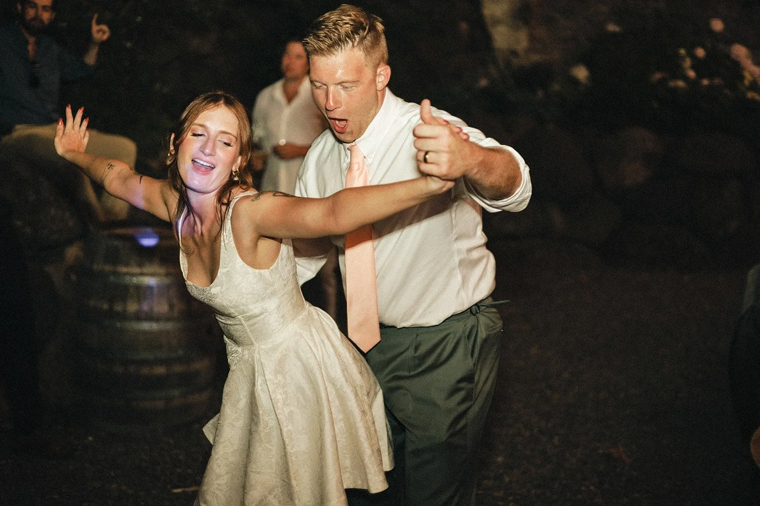 A man and woman dancing closely together at a celebration, with the woman wearing a light-colored dress and the man wearing a white shirt and tie, in a dark indoor setting.