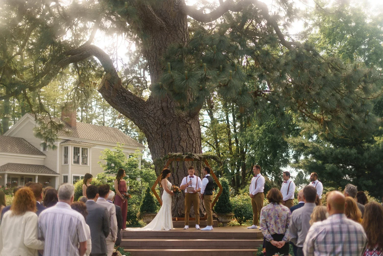 Outdoor wedding ceremony under a large tree with a wooden platform. The bride in a white dress and the groom in a white shirt with suspenders are standing at the center, with officiant reading from a paper. Bridesmaids and groomsmen are standing on either side. Guests are seated and standing, watching the ceremony, in front of a white house with a large porch.