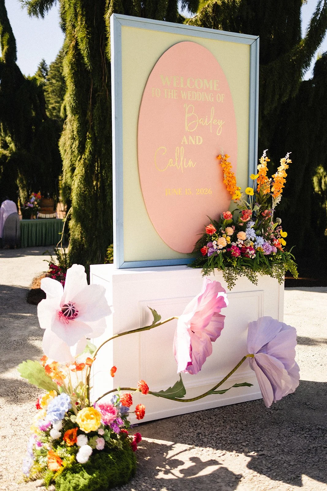 Wedding welcome sign with pink and yellow design, floral arrangements, and outdoor setting.