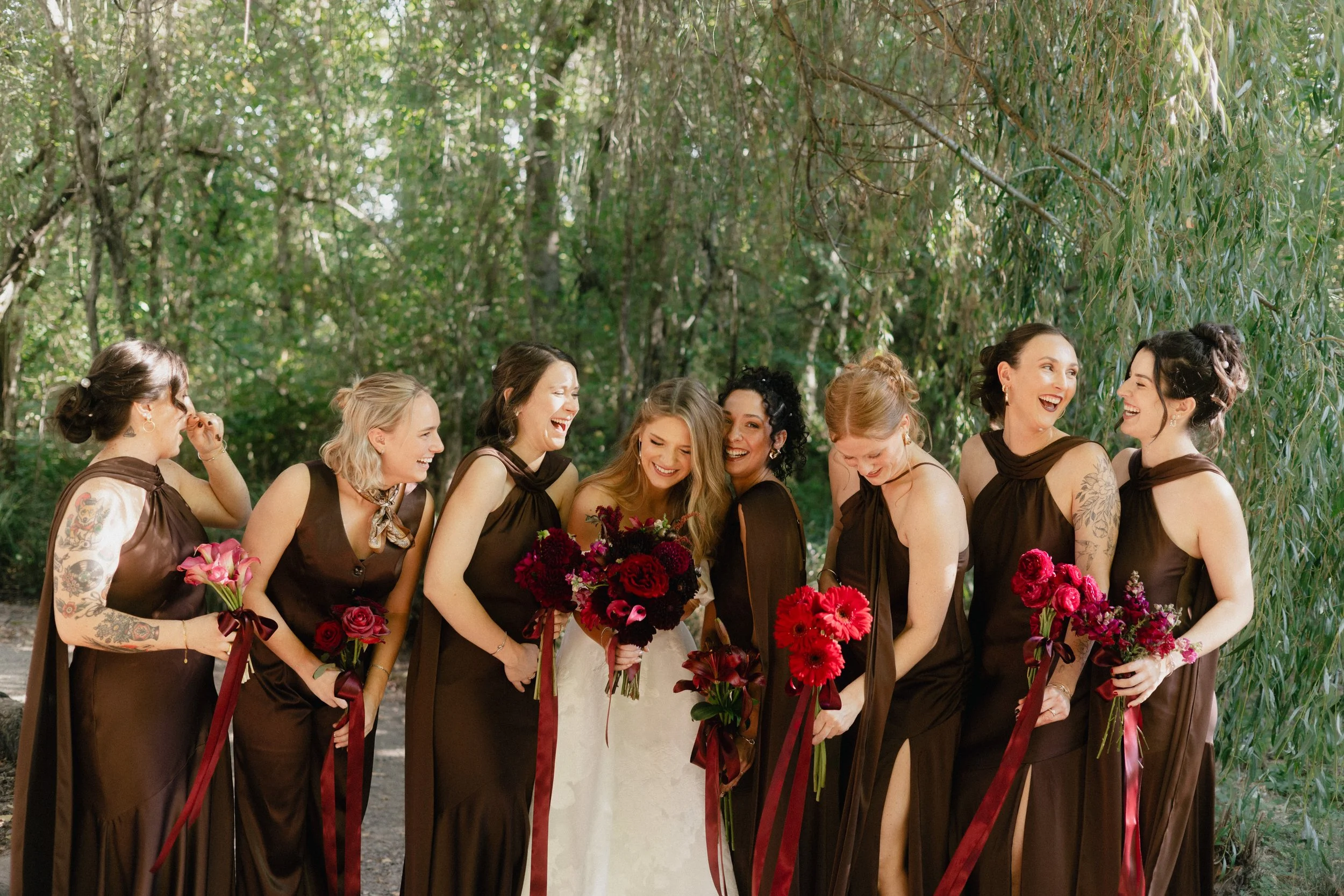 A group of women dressed in matching brown dresses, holding bouquets, and laughing together outdoors in a wooded area.