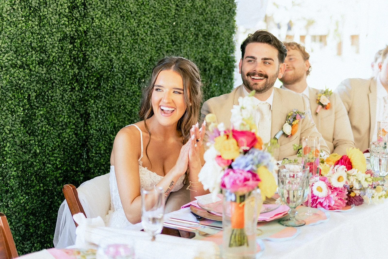 A group of people at a wedding reception, sitting at a decorated table, smiling and enjoying the celebration.