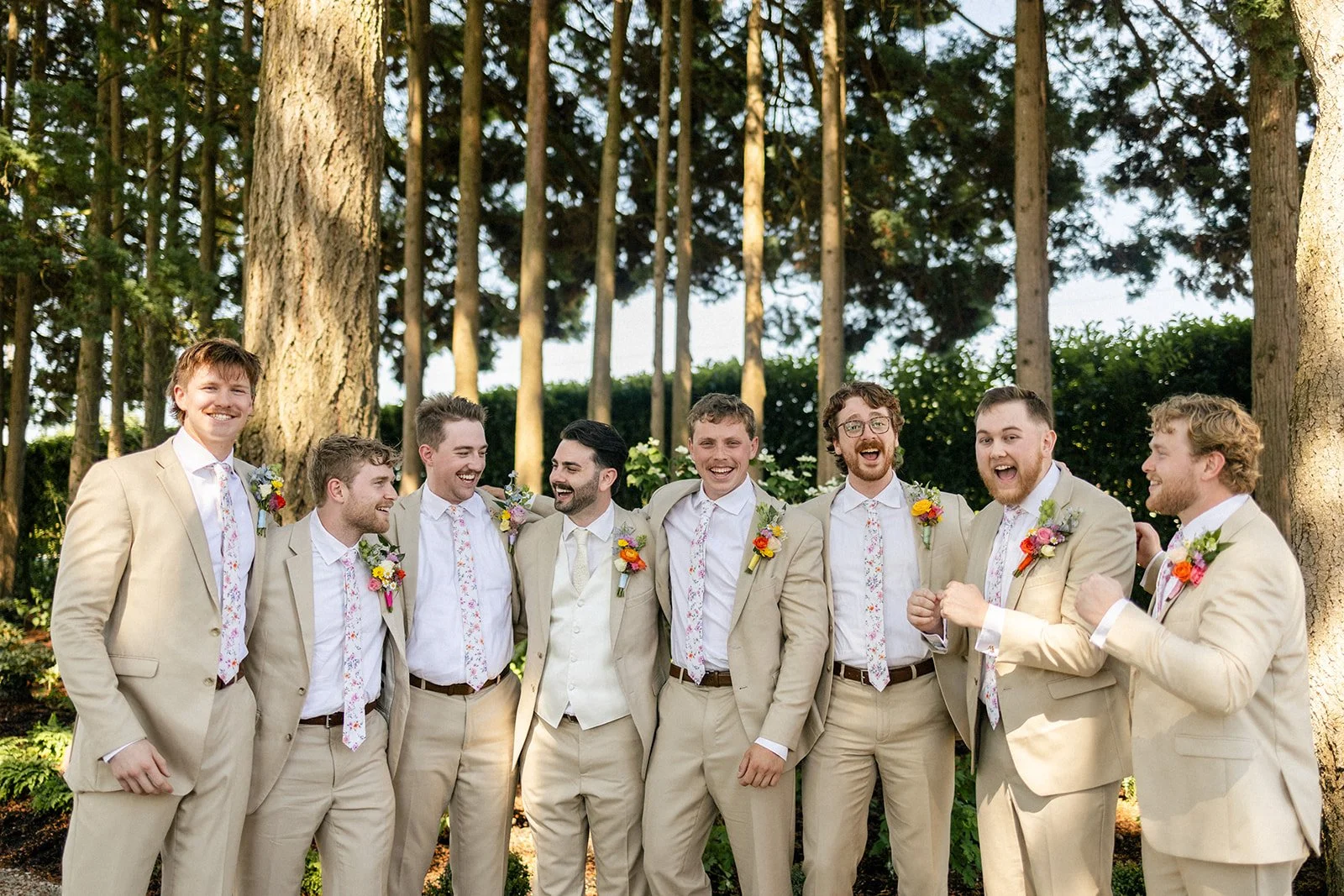 A group of nine men in beige suits and floral ties standing outdoors with trees in the background, smiling and laughing together.
