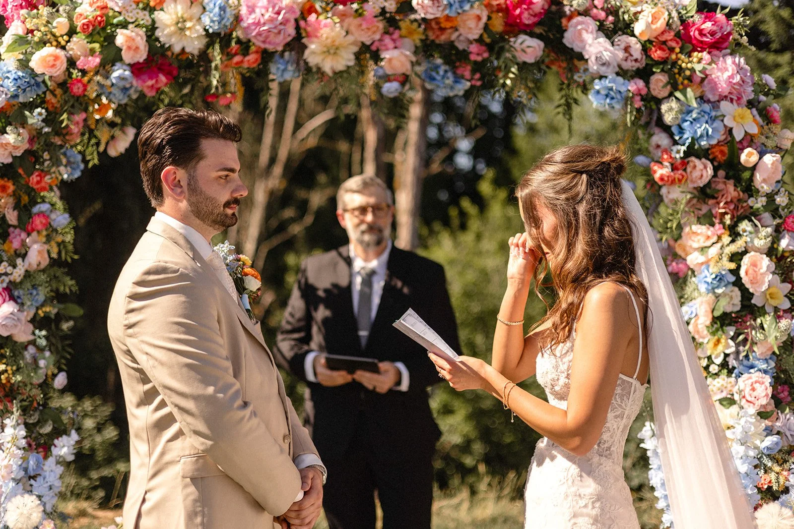 A bride and groom exchanging vows at an outdoor wedding ceremony, standing under a floral arch with an officiant in the background.