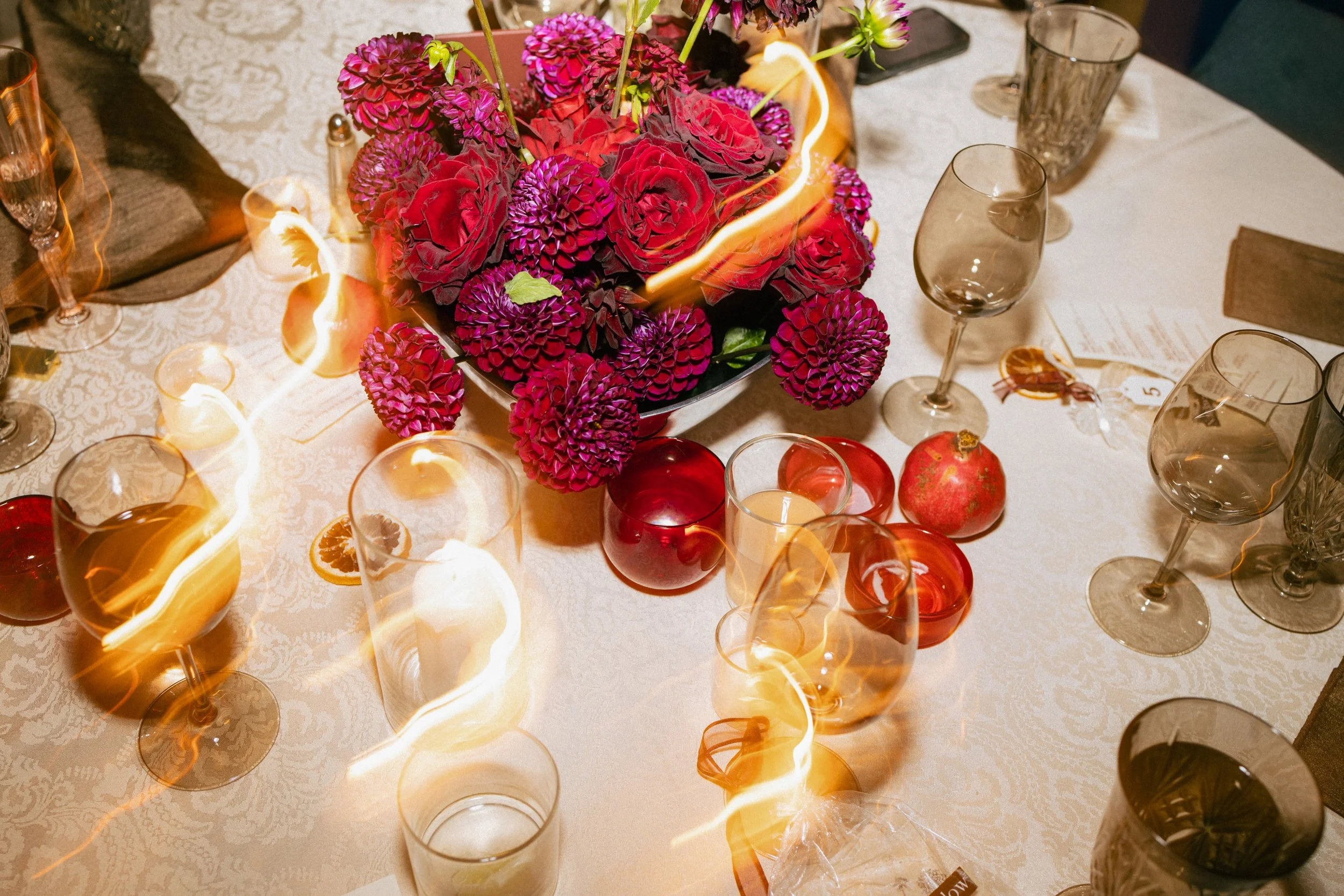 A table decorated with pink and purple flowers as a centerpiece, surrounded by empty wine glasses, candles, and a pomegranate, on a white tablecloth.