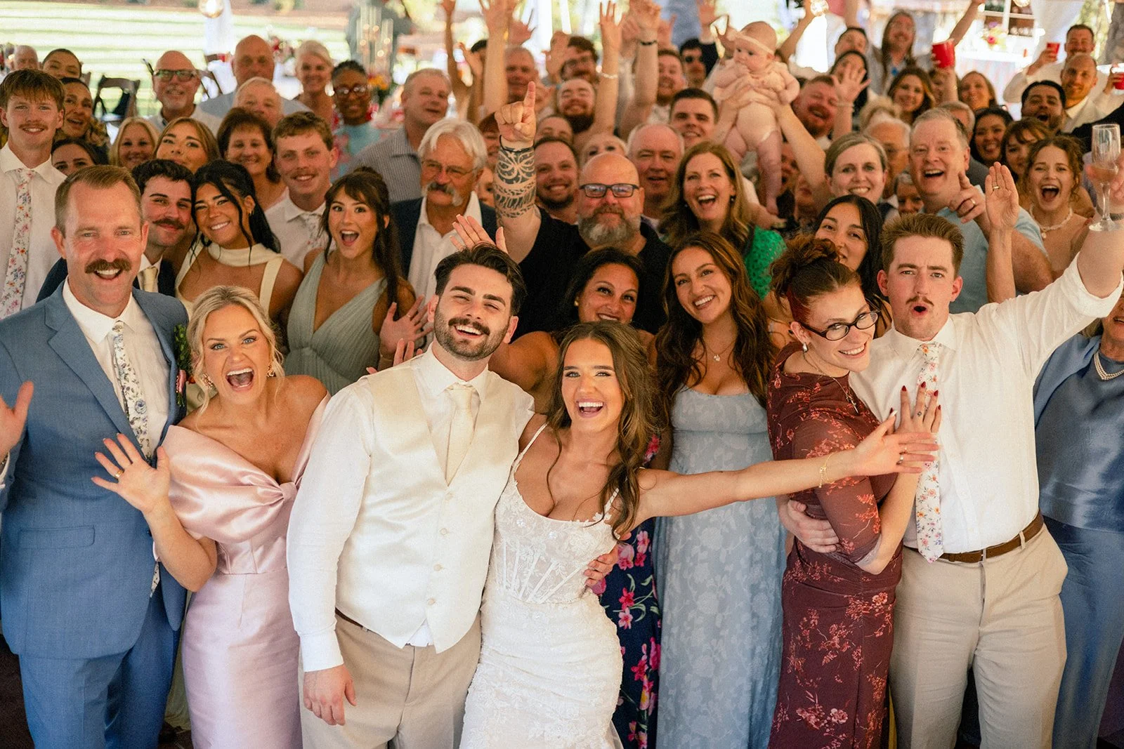 A large group of smiling people at a wedding celebration, including the bride and groom in the front center, celebrating together.