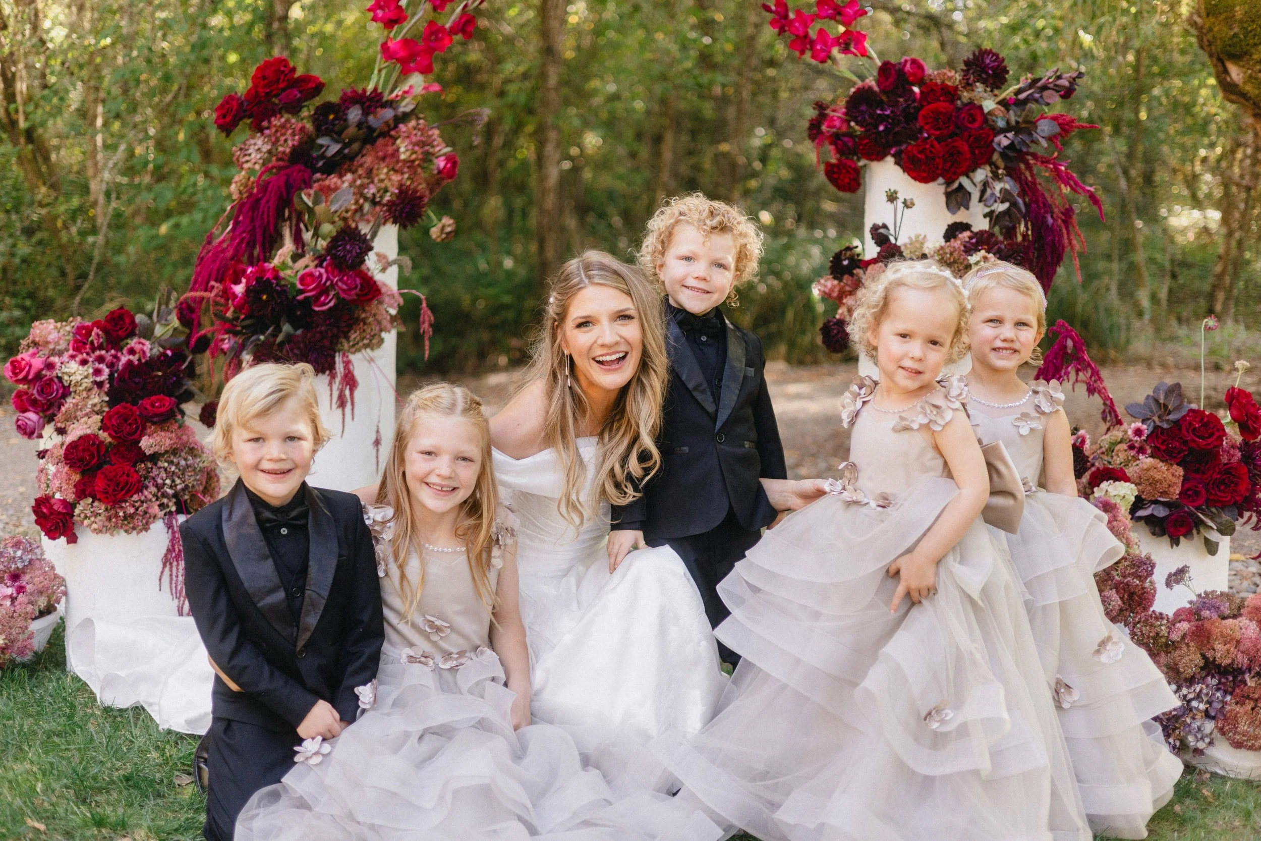 Children and a woman in formal dresses outside, posing under a floral arch for a photo.