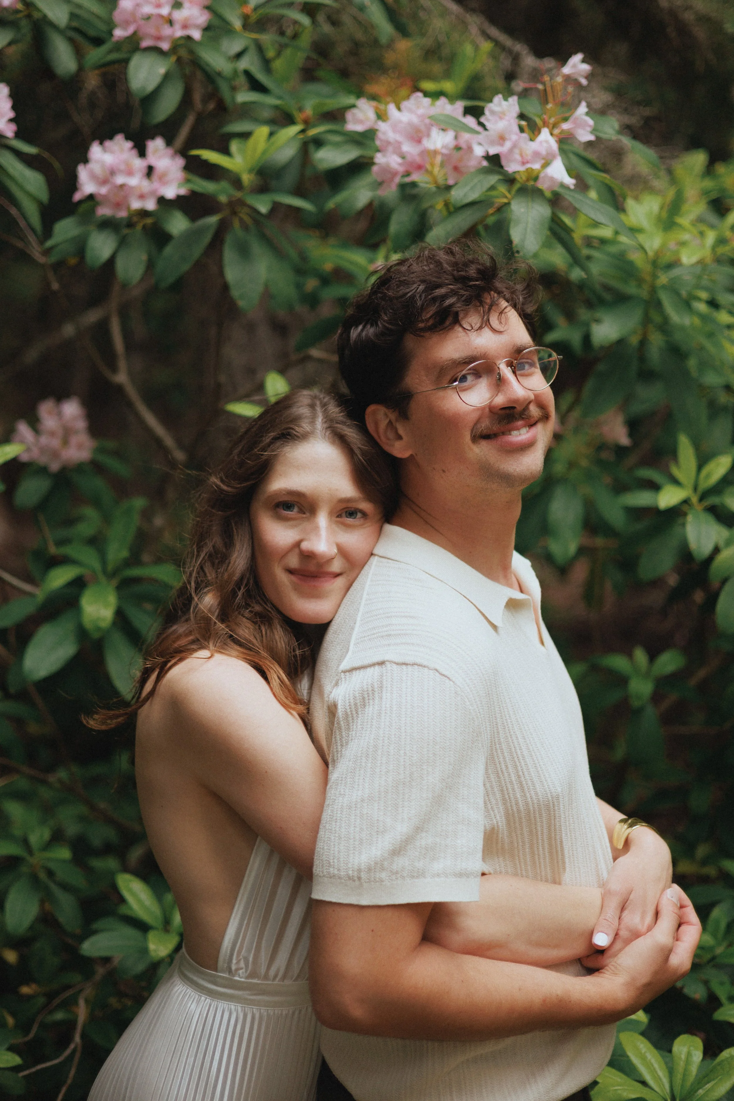 A young woman with long wavy brown hair and fair skin is hugging a man from behind. She is smiling softly with her eyes open and is wearing a sleeveless, light-colored dress. The man has curly brown hair, glasses, light skin, and is smiling with his teeth showing, wearing a light-colored, short-sleeved polo shirt. They are standing outdoors in front of lush green foliage with pink rhododendron flowers.