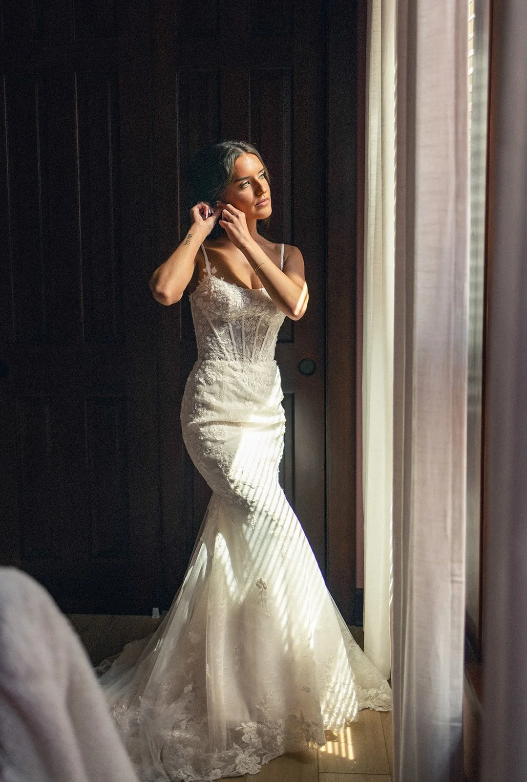A woman standing by a window, adjusting earrings, in a sleeveless lace and satin wedding dress with a mermaid silhouette.