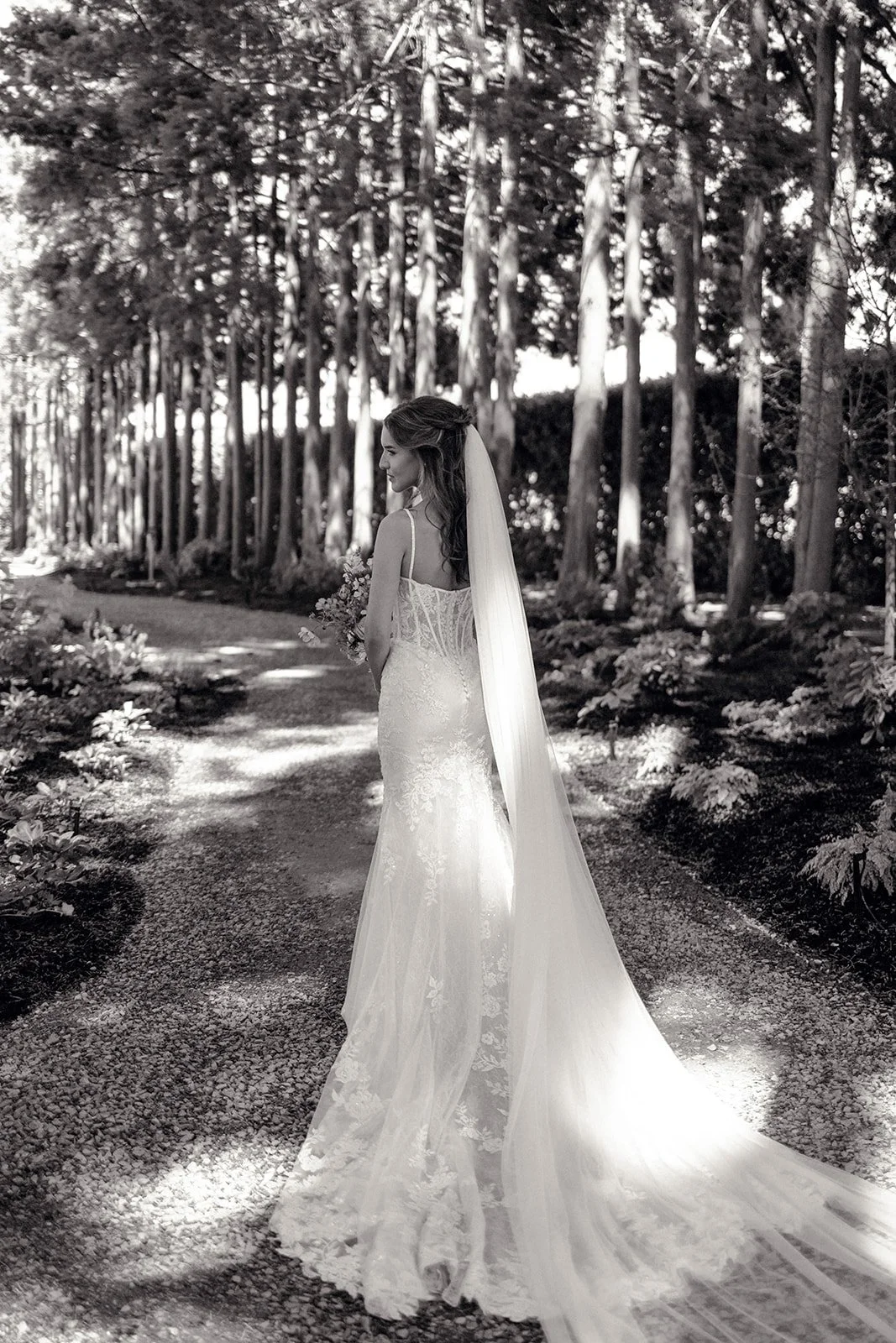 A bride in a lace wedding dress and veil standing on a forest path surrounded by trees and flowers.