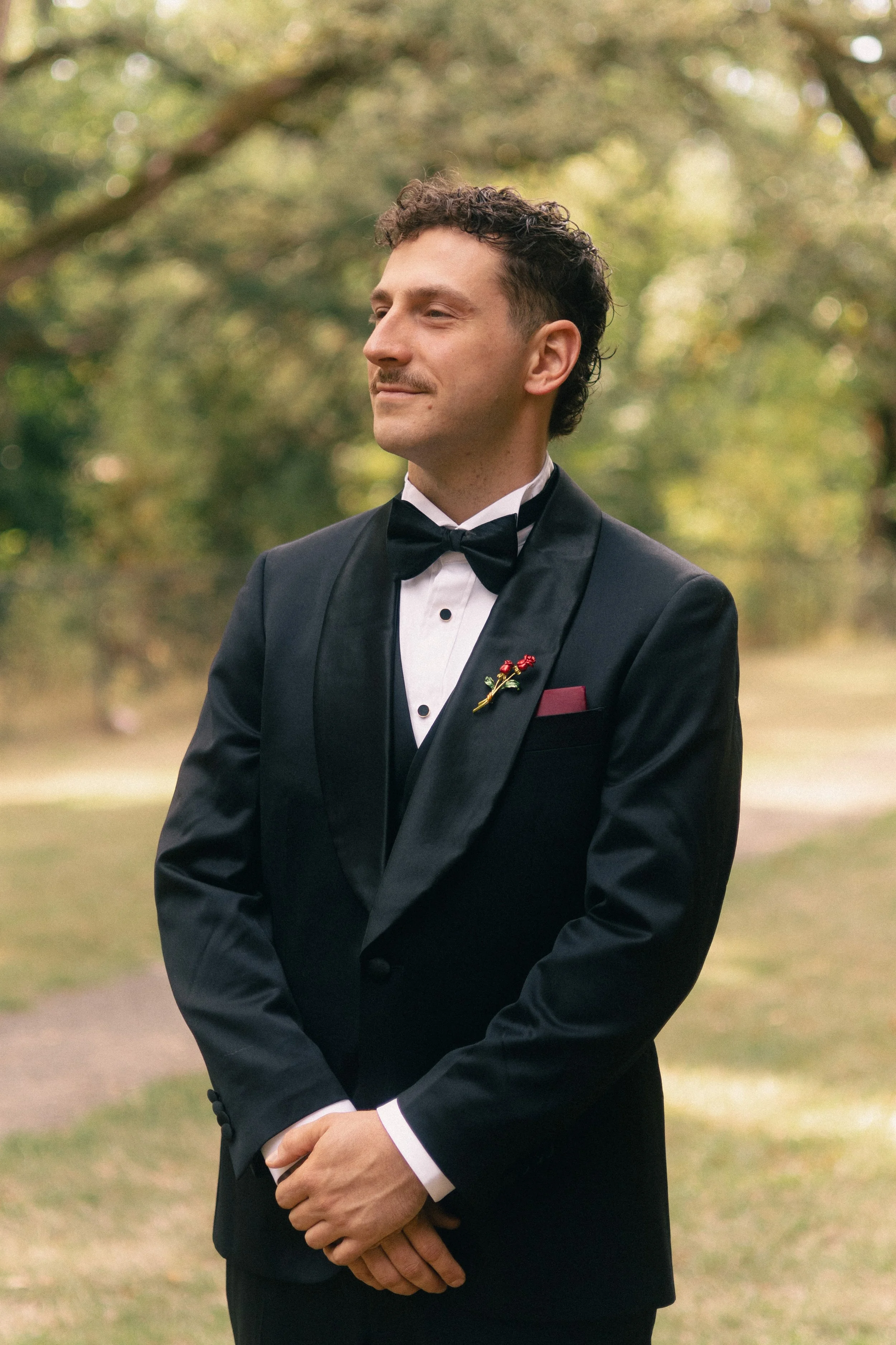 A man dressed in a black tuxedo with a bow tie, standing outdoors in a park-like setting with blurred trees in the background. He has dark, curly hair and a slight smile.