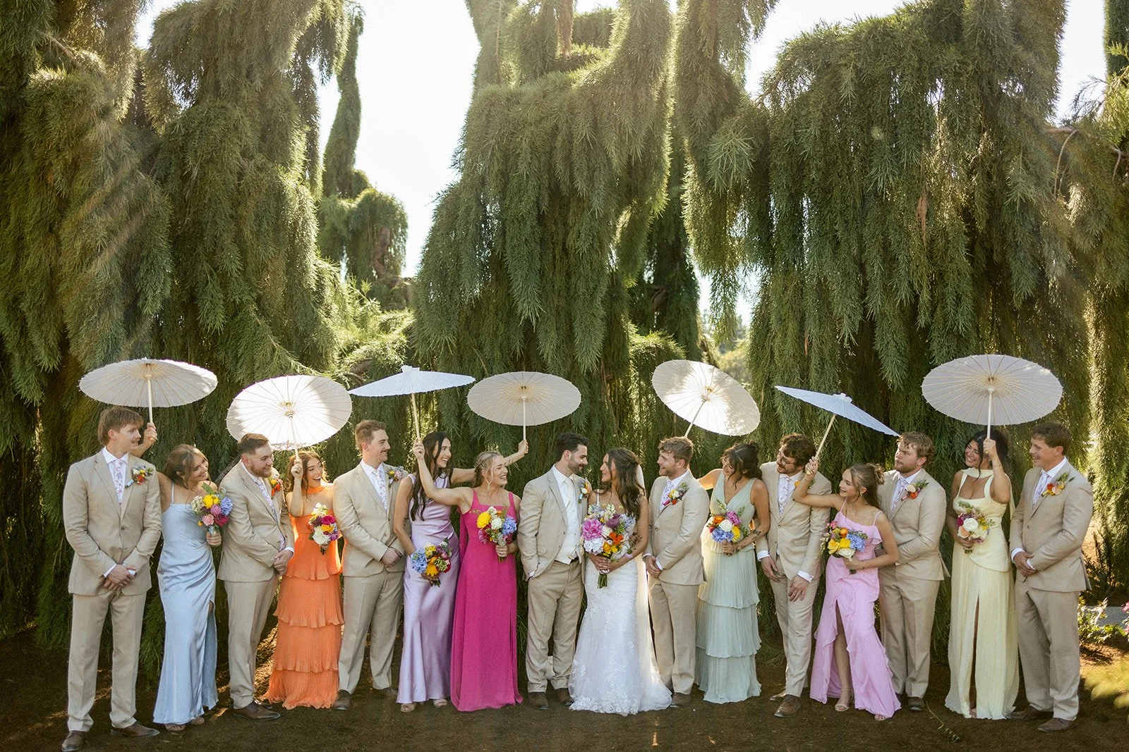 A wedding party of 14 people, including the bride and groom, standing outdoors in front of tall trees. The bride is dressed in a white gown, and the groom and the men are in beige suits. The women are wearing colorful dresses in pastel shades, and mo
