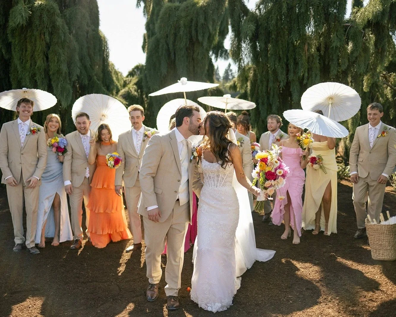 A wedding scene outdoors with a bride and groom kissing in the center, surrounded by bridesmaids and groomsmen holding white parasols and bouquets, under a clear sky with tall trees in the background.