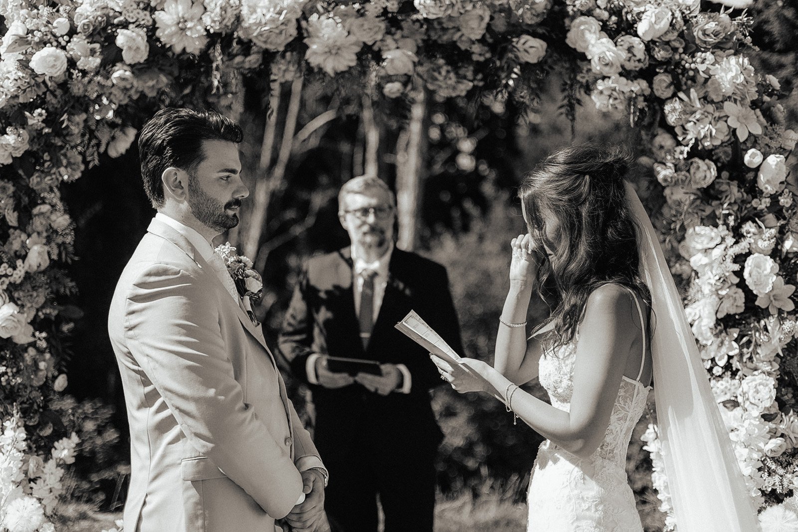 A bride and groom exchanging vows in a wedding ceremony outdoors at night, under a floral arch, with an officiant standing behind them. The bride is wiping tears with her hand, and both are dressed in formal wedding attire.