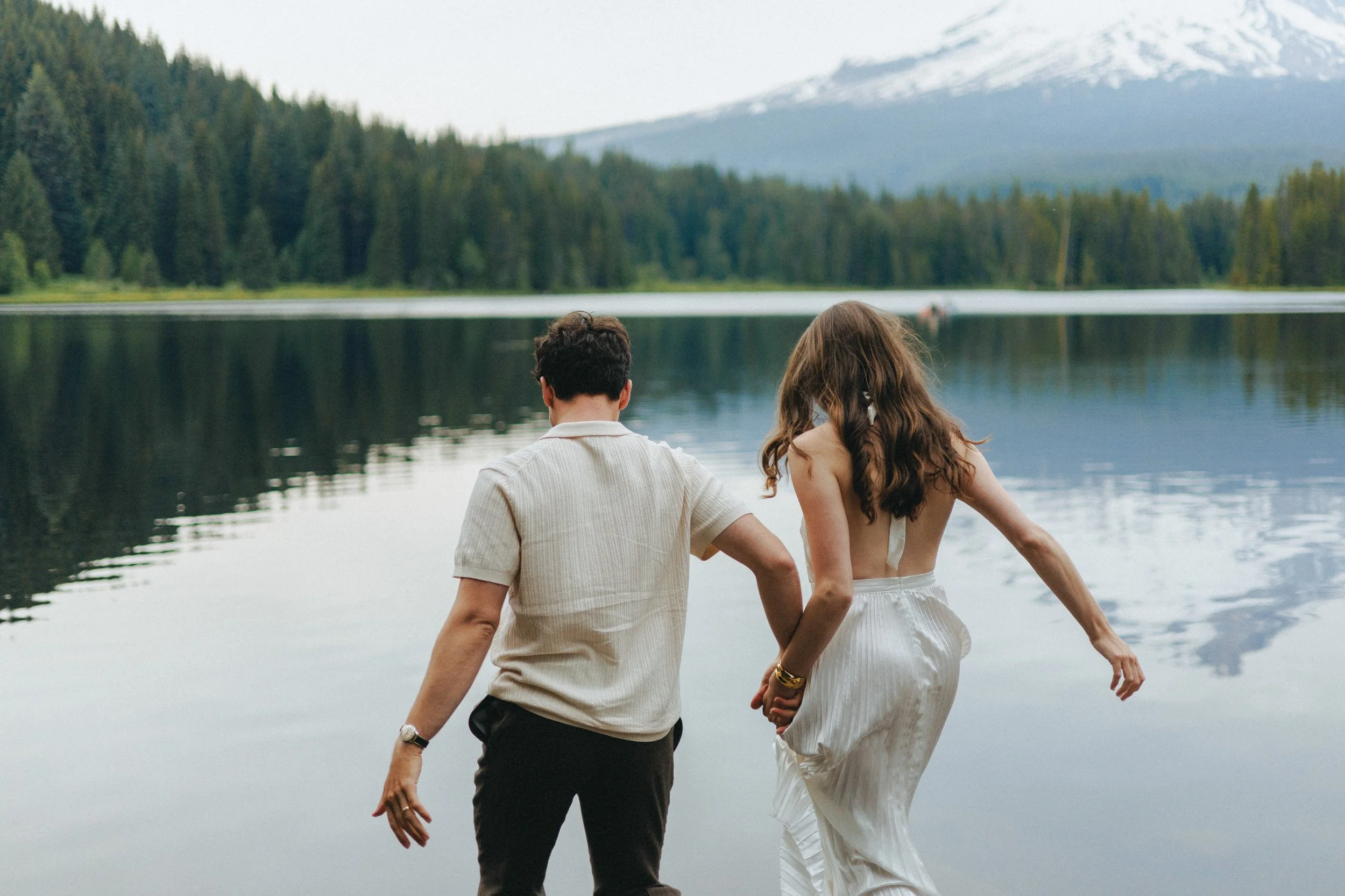 A couple holding hands walking by a lake with a mountain and forest in the background.