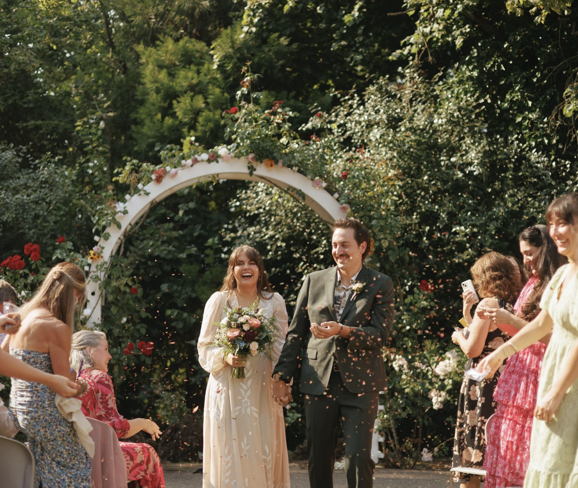 A couple at their outdoor wedding ceremony, holding hands and smiling as confetti falls around them, with friends and family celebrating.