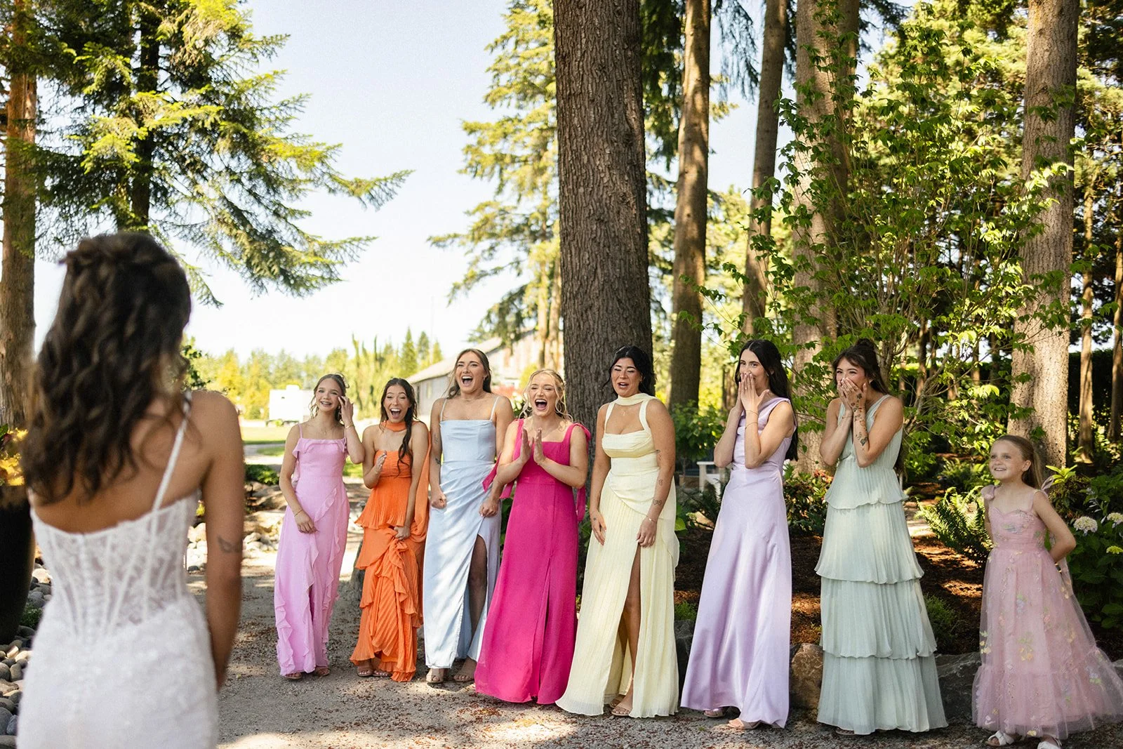 A woman in a white dress with her back to the camera is looking at eight women, of various ages, wearing colorful dresses, standing outdoors among tall trees. They are smiling, laughing, and covering their mouths in surprise or joy.