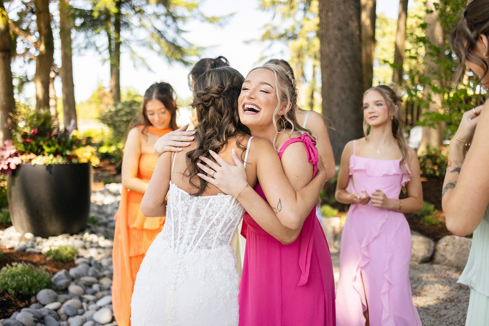 Two women hugging at an outdoor wedding, surrounded by flower arrangements and other women in colorful dresses, with trees in the background.