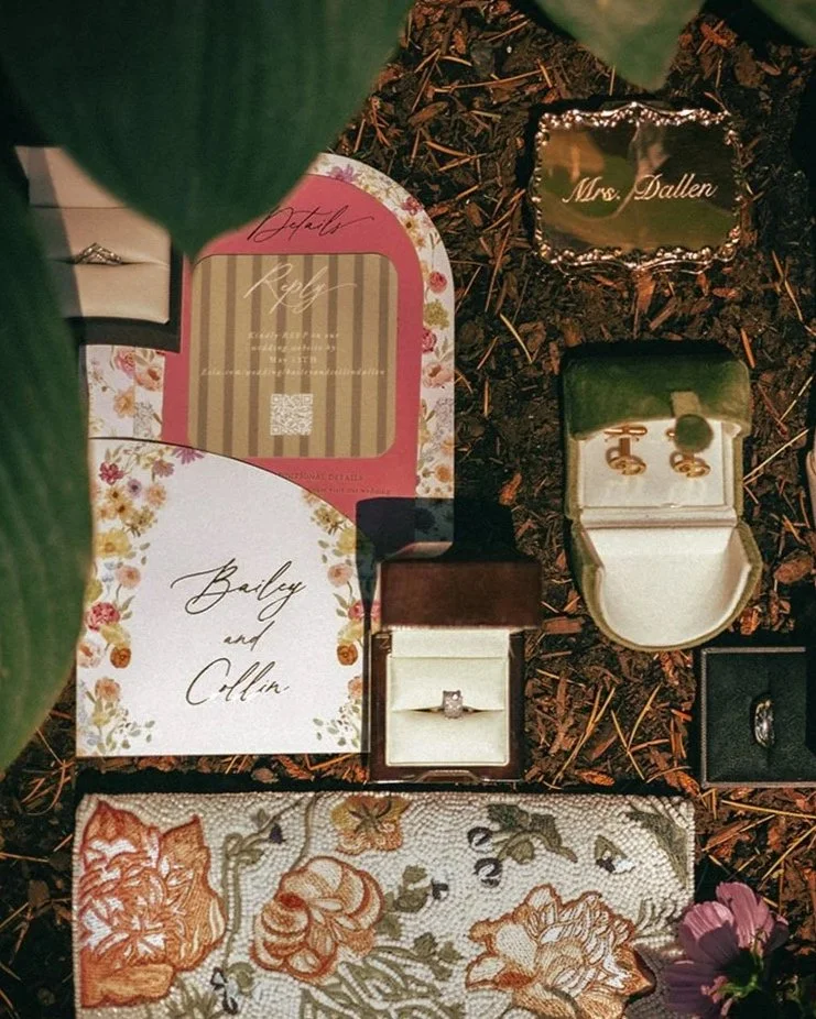 A display of wedding accessories and stationery on the ground, including an engagement ring, earrings, wedding bands, and invitations, decorated with flowers and labeled 'Mrs. Dallen' and 'Bailey and Collin'.
