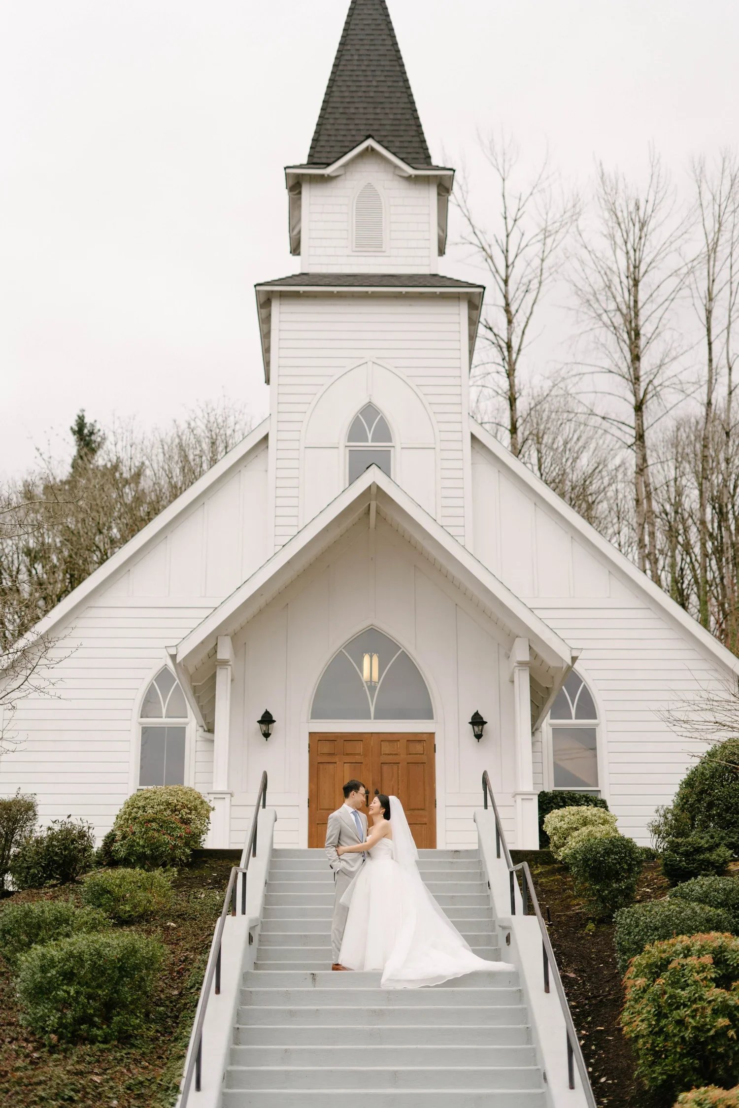 A bride and groom embrace on the steps of a white church in this charming Portland wedding scene.