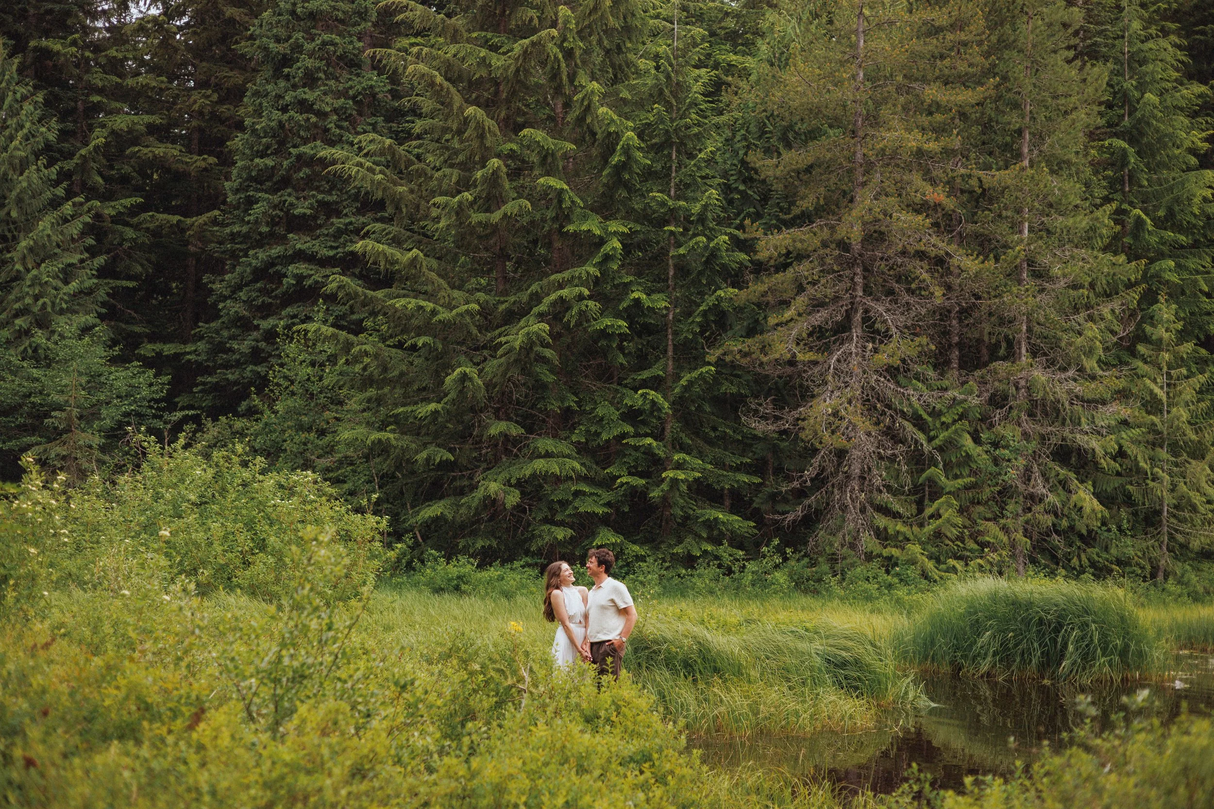 A couple stands holding hands beside a small pond surrounded by lush greenery and tall pine trees in a forest.