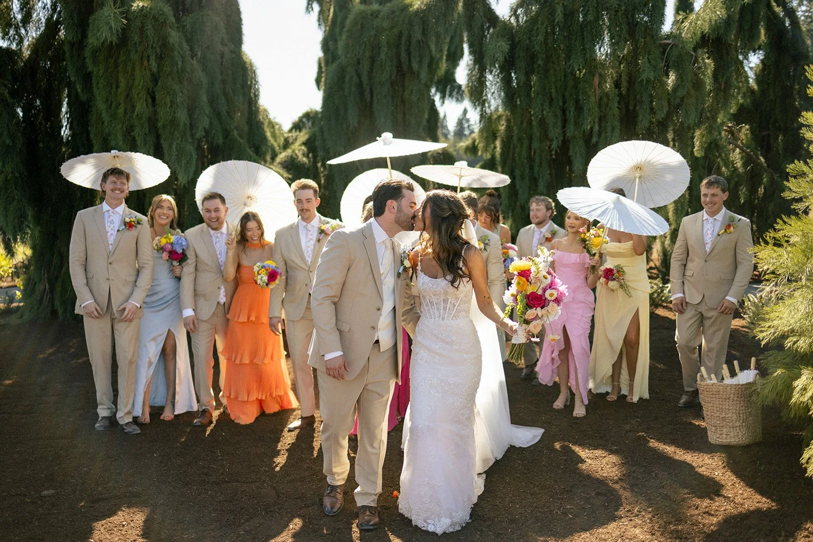 A wedding ceremony outdoors with newlyweds kissing, surrounded by wedding party holding umbrellas and bouquets under tall green trees.