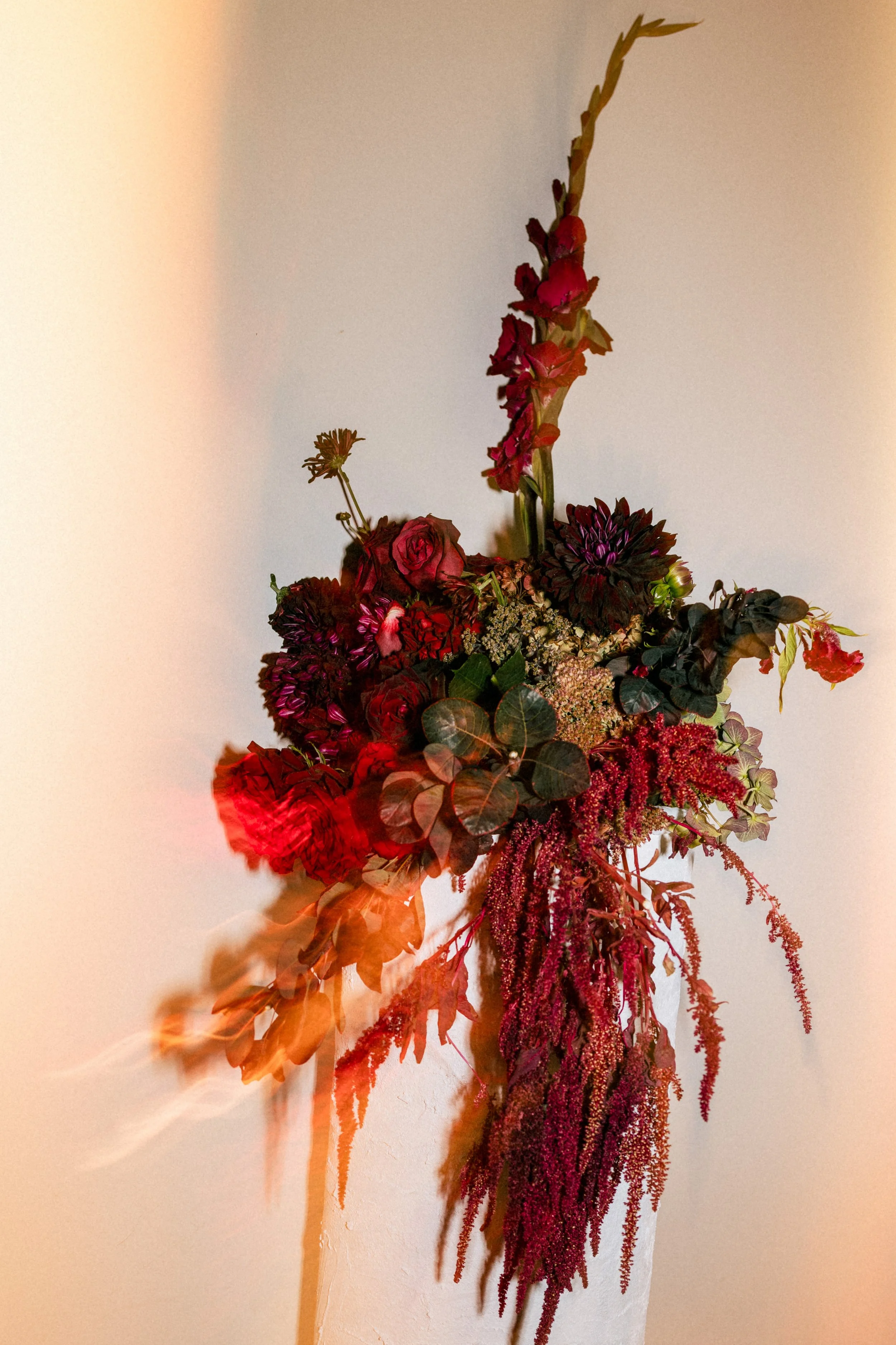 A wall-mounted floral arrangement featuring various red and dark purple flowers and green foliage, with a vertical element extending upward.